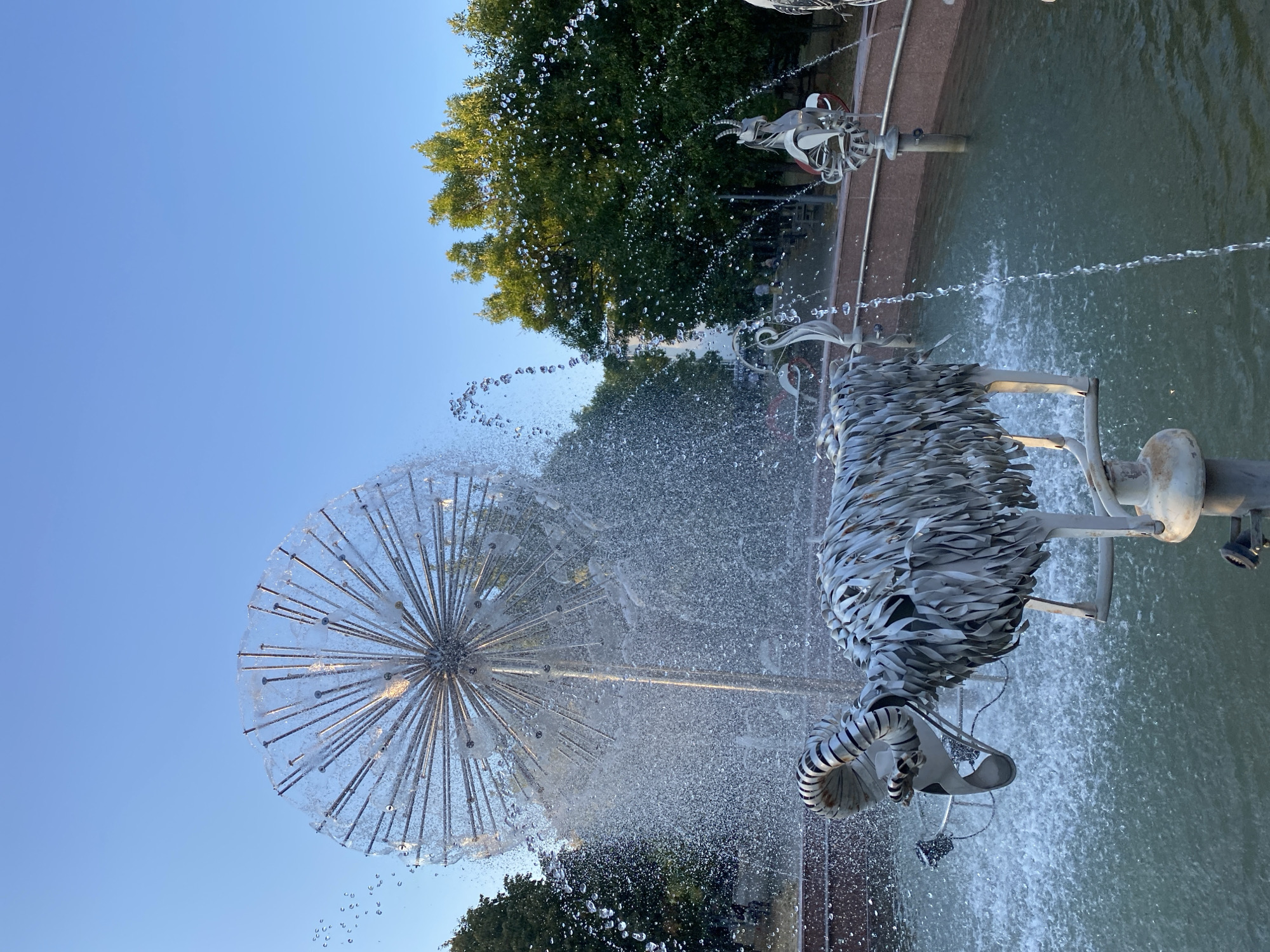The image shows a fountain with a large, spherical sculpture resembling a dandelion clock, with thin metal rods radiating outwards. Water sprays from this sculpture, creating a misty effect. In the foreground, there is a metal sculpture of a ram with large, curved horns. The ram's body is made of many thin, overlapping metal strips, resembling fur. The fountain contains water with green algae or sediment. In the background, there are trees with lush green foliage and a clear blue sky. To the right, there are other metal sculptures of animals in the fountain, including what appears to be a goat. This fountain is located in the Alley of Classics in Chișinău, Moldova, a public park featuring sculptures of prominent Moldovan writers and poets, often surrounded by decorative elements like fountains and gardens.