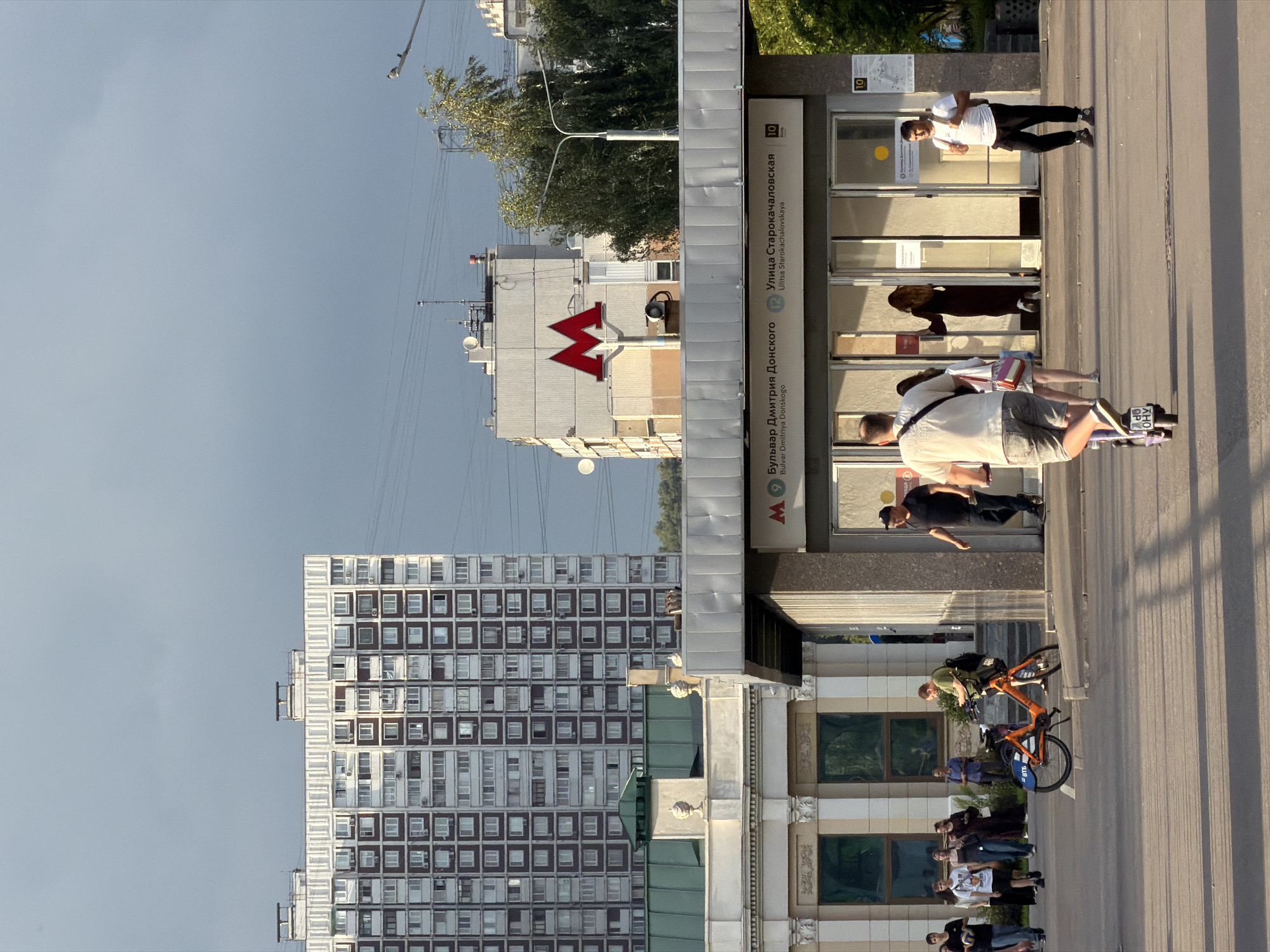 The image captures an outdoor scene in Moscow, Russia, with a focus on a metro station entrance. In the background, a tall, white residential building with many windows dominates the skyline under a cloudy, grey sky. To the right of this building is another, less tall building with a large red 'M' logo, signifying the metro. The foreground shows the entrance to the metro station, which has glass doors and signage indicating "Bulvar Dmitriya Donskogo" and "Ulitsa Starokachalovskaya." Several people are visible around the entrance and on the surrounding paved area. One man is riding a scooter away from the viewer, while another man walks towards the metro entrance carrying a backpack. Other individuals are standing or walking near the station. An orange shared bicycle is parked on the sidewalk to the left. The overall atmosphere suggests a typical urban day, with people commuting and utilizing public transportation.
