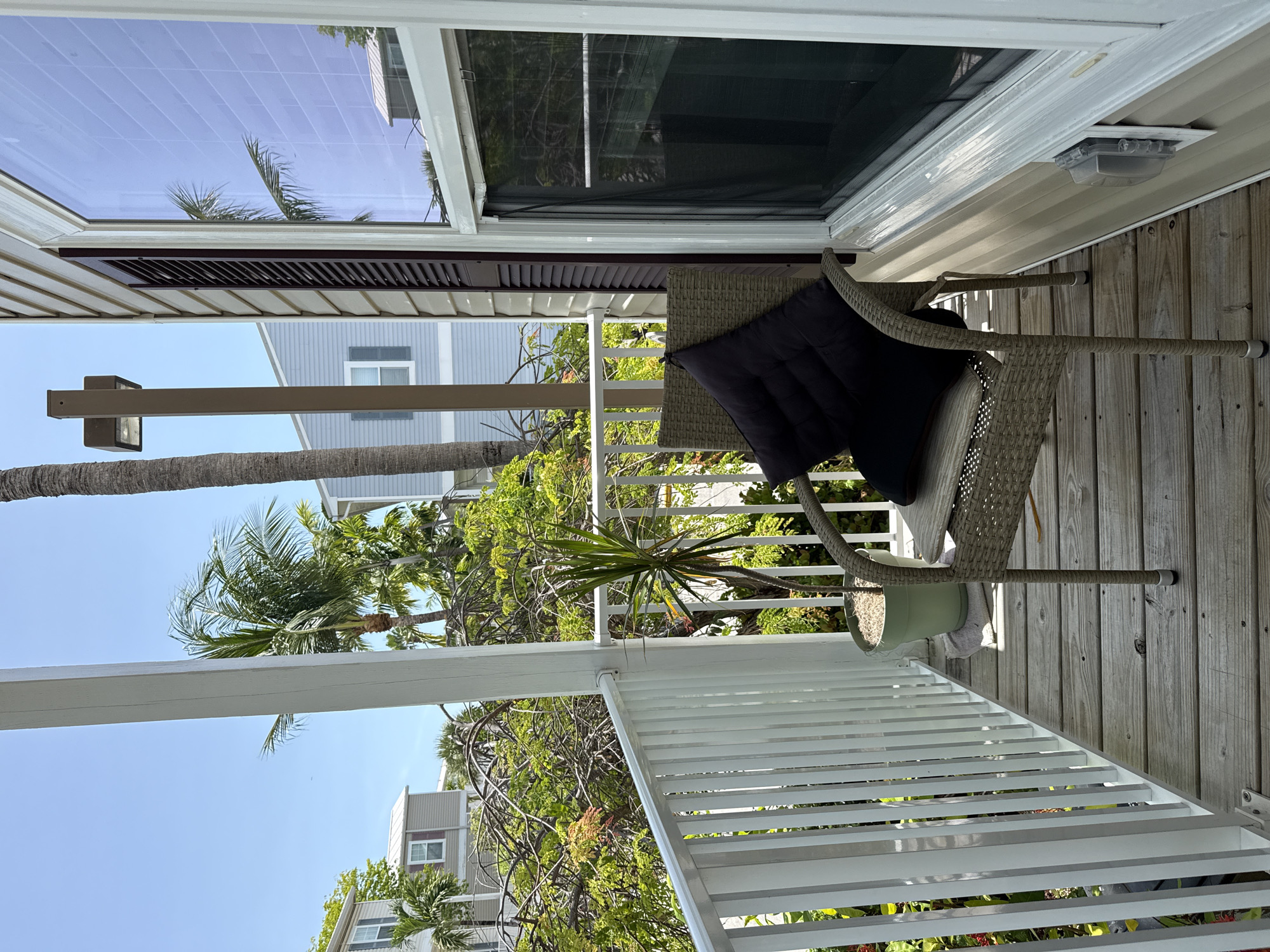 A woven wicker chair with a black cushion sits on a wooden deck, facing away from the camera. The chair is positioned on a porch with a white railing to the left, which separates the deck from a backdrop of lush green trees and a bright blue sky. In the background, two-story houses with light blue siding and white trim are visible, interspersed with palm trees. A tall, slender outdoor light fixture stands to the right of the chair, and a large window with a white frame is situated on the right side of the image. The overall scene suggests an outdoor residential setting on a sunny day.