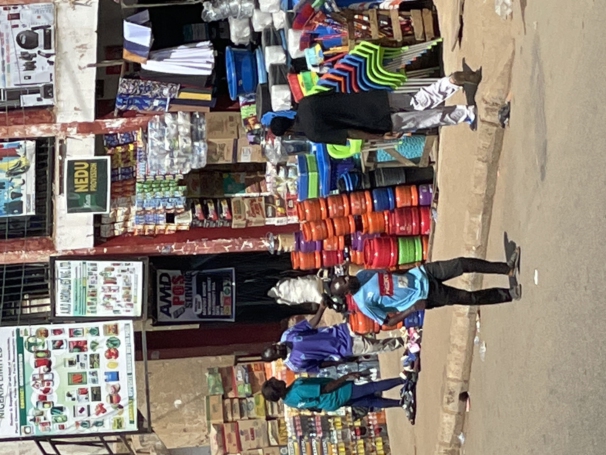 The image depicts a street scene in Bukuru, Nigeria. A roadside shop displays a variety of goods, including plastic containers, chairs, and packaged food items. Several people are present. One man wears a black shirt and white pants, another wears a blue shirt and dark pants. Two other people wear blue and green shirts. The shop's signage includes "NEDU PROVISION" and "AMD POS SERVICES." Additional advertising posters are affixed to the building's facade. The street surface is visible in the foreground.