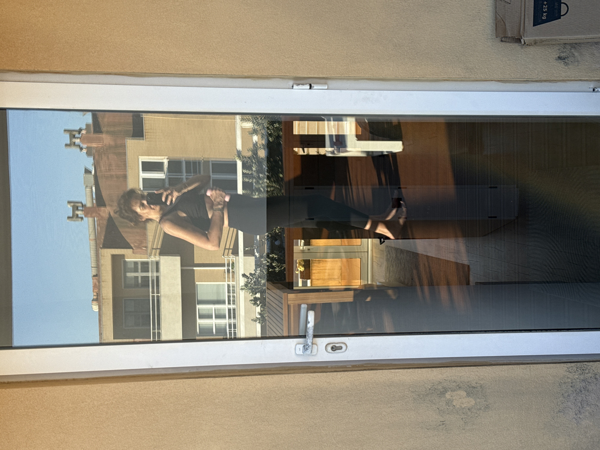 The image shows the reflection in a window of a person holding a mobile phone, likely taking a photo or video. The person appears to be standing on a balcony. Reflected in the window are buildings, indicating an urban environment. The location is Vila Nova de Gaia, Portugal, based on architectural style and environment. Other reflected objects include patio furniture and plants on the balcony. A cardboard box is visible in the upper right corner of the frame. The window frame is white.