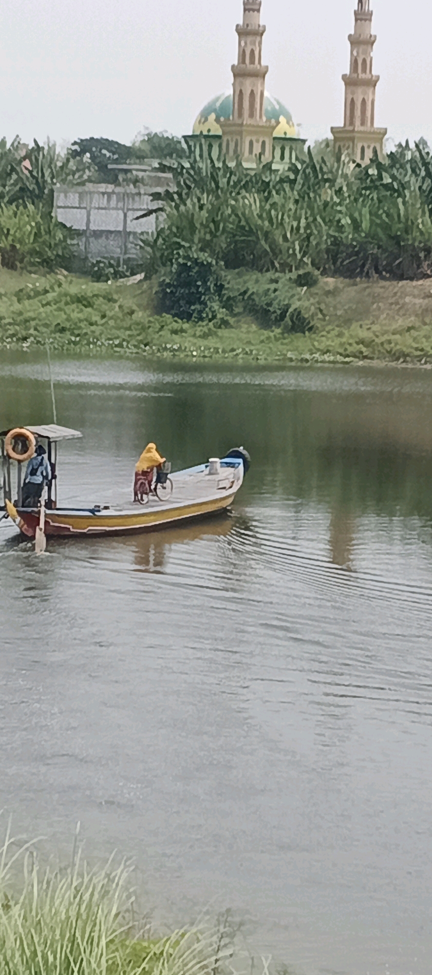 In Brumbun, Indonesia, a small motorized boat with a yellow hull and white superstructure navigates a body of water. A person wearing a yellow garment is seated on a bicycle positioned on the boat's deck, facing forward. Another individual, dressed in a blue shirt and dark pants, stands near the front of the boat, holding onto a railing. A life preserver, orange in color, is attached to the boat's canopy. The water is calm with visible ripples. In the background, lush green vegetation lines the far bank of the water, and a mosque with two prominent minarets and a dome is visible above the trees. The sky is overcast.