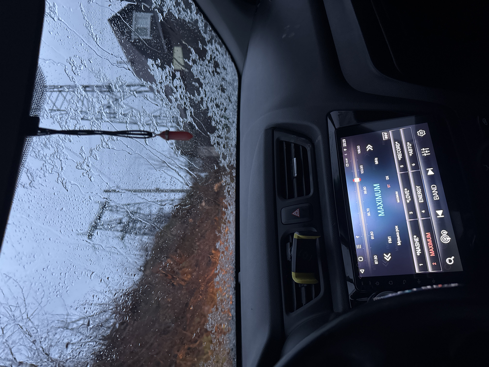 The image, taken from inside a vehicle, shows a rainy windshield with visibility obscured by water droplets. Through the windshield, a blurred landscape is visible, including a building and power lines, suggesting an urban or suburban environment. Inside the vehicle, the dashboard is visible, featuring air vents, a hazard light button, and a modern infotainment system displaying a radio interface tuned to a station called "MAXIMUM" and other FM stations. Based on the "Музыка рок" ("Music Rock") label and the FM frequencies displayed, it's possible the location is Chaikovsky, Russia.