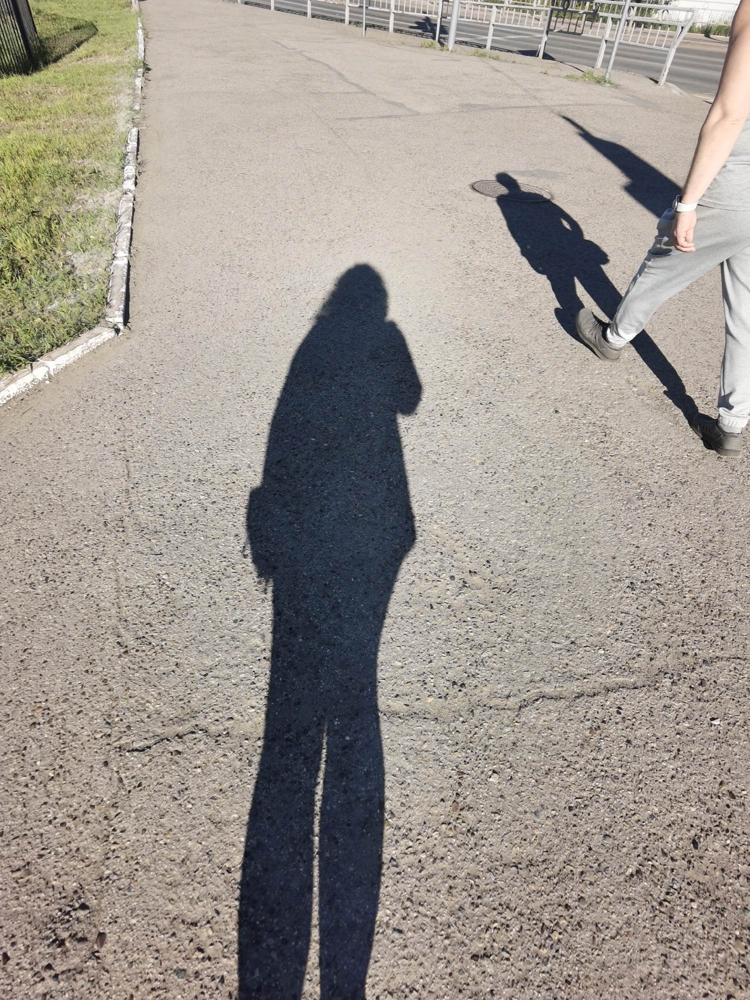 The image captures a daytime scene on a paved pathway in Chaikovsky, Russia. Two distinct shadows are visible on the rough asphalt surface. The larger shadow, cast by a person standing closer to the camera, stretches across the foreground. This shadow is elongated and appears to be in a relaxed stance, possibly holding a phone or camera.
A second, smaller shadow is cast by a person walking further down the pathway, towards a manhole cover. This shadow is angled and shows the person's legs in motion. The individual casting this shadow is wearing light-colored sweatpants and dark shoes. In the background, a metal fence runs parallel to the pathway, and beyond that, a road and some vegetation are discernible. The overall environment suggests a public outdoor space, possibly a park or a pedestrian walkway within the city.