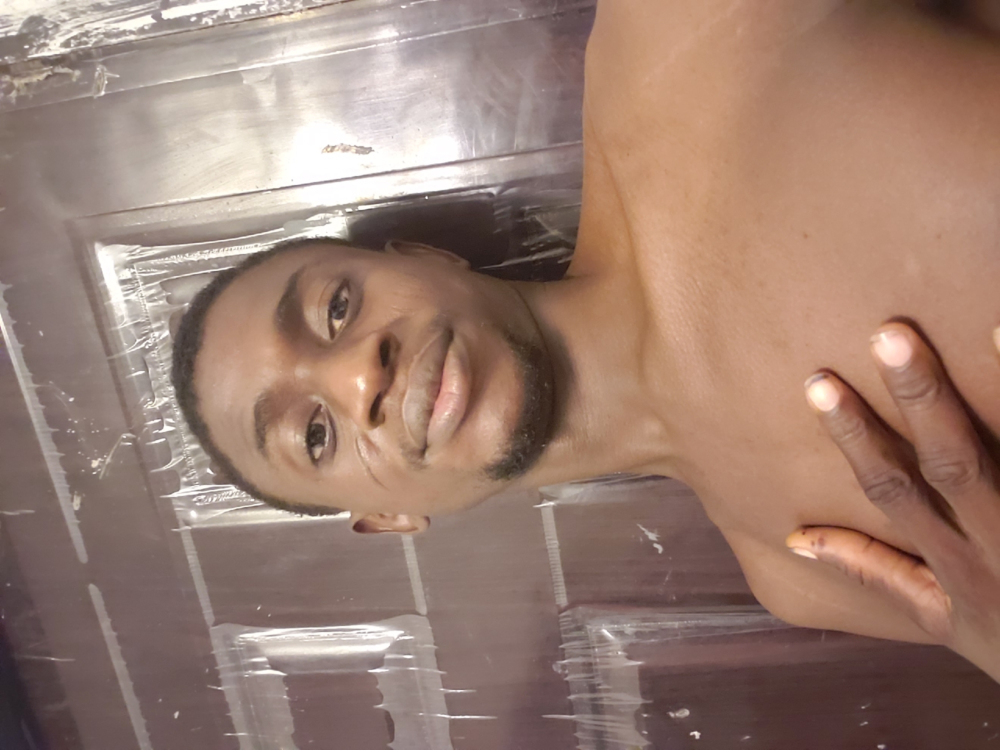 A medium shot depicts a shirtless dark-skinned man with a trimmed beard, lying on his side on what appears to be a shiny, reflective surface with linear patterns, possibly plastic covering a door or table. His right hand rests on his shoulder. The location is not discernible as Adonkolo, Nigeria, based solely on visual information.