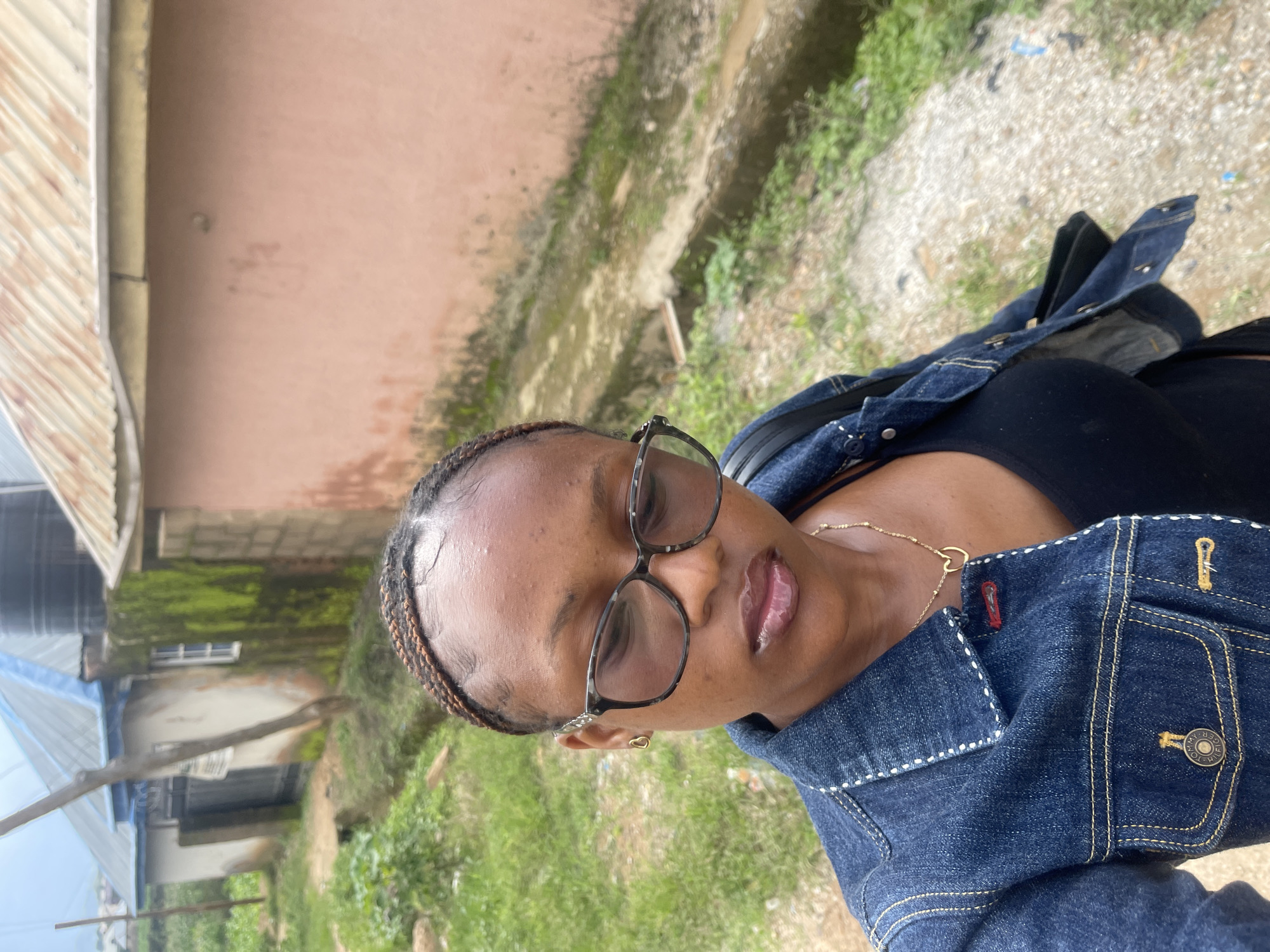 A medium shot captures a young Black woman wearing a denim jacket and a black tank top, with a gold heart-shaped necklace. She has dark braided hair and is wearing glasses with a tortoise-shell pattern. The woman is looking down, her lips slightly parted. The background shows a rustic environment with buildings and overgrown vegetation, suggesting a location in Rafin Par, Nigeria. The buildings appear to be made of concrete and brick, with corrugated metal roofs. The ground is a mixture of dirt and gravel, with evidence of a drainage ditch. The overall impression is of a casual, outdoor setting in a rural or semi-rural area.