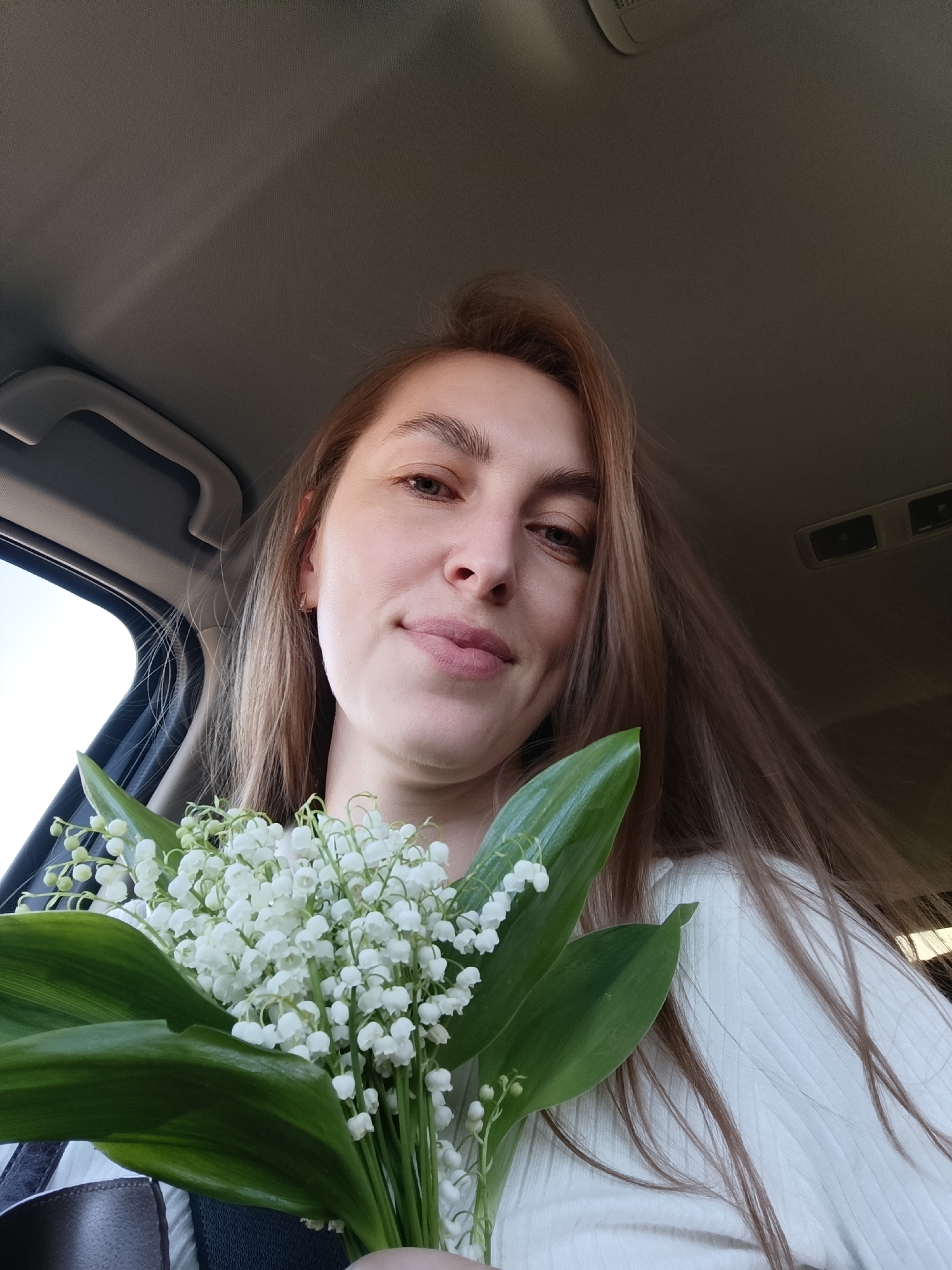 A young woman is taking a selfie in what appears to be a car. She is holding a bouquet of lily of the valley flowers. The interior of the car is visible, including the ceiling, a window, and a seatbelt. The location cannot be determined from the image alone.