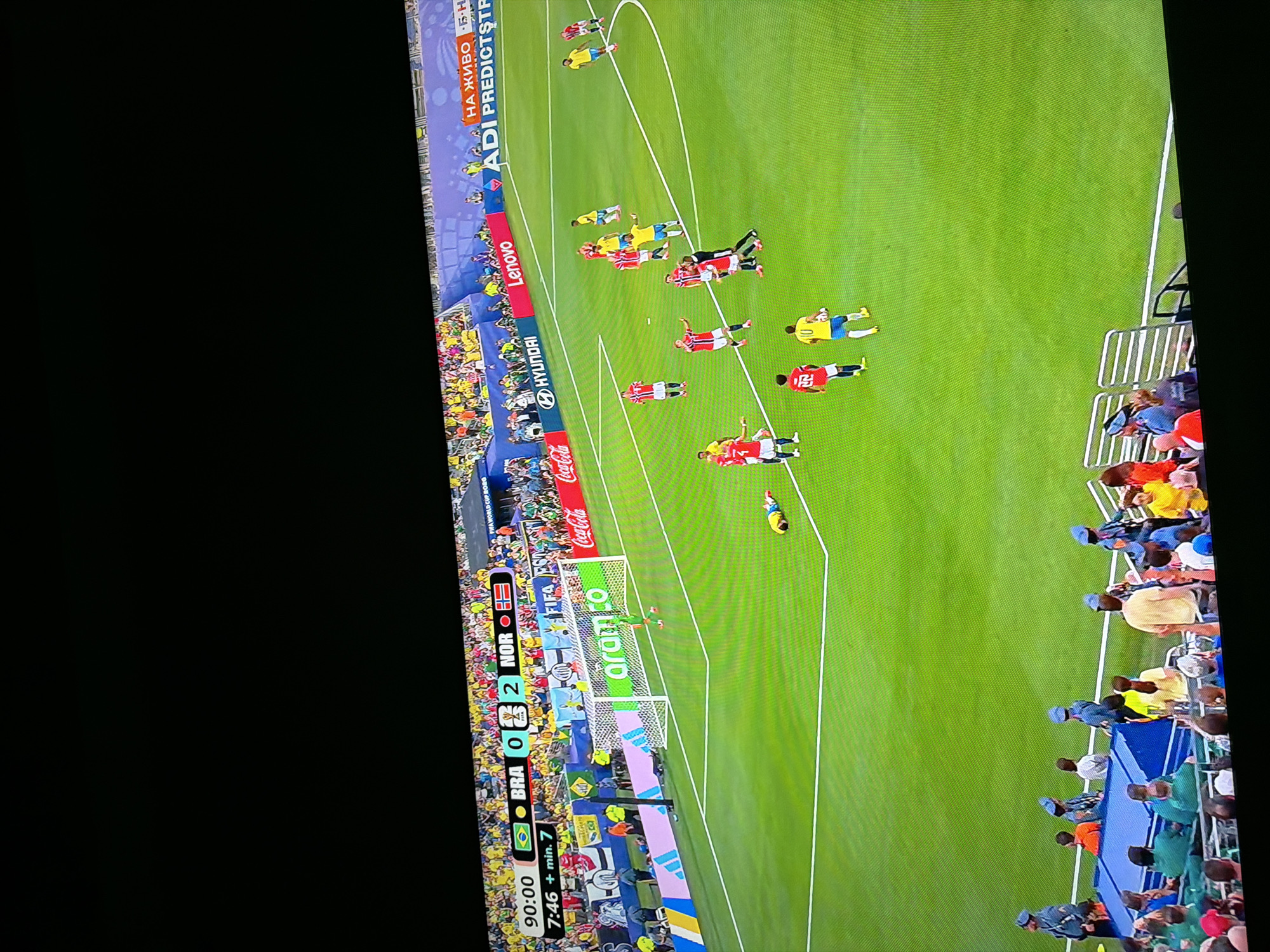 The image shows a soccer match on a green field under stadium lights. A scoreboard at the top left indicates the time as 90:00 and the score as BRA 0, NOR 2, with approximately 7:46 minutes of added time. The players are wearing red and yellow uniforms. In the foreground, spectators are visible in the stands. The scene is likely a professional soccer match being broadcast. There is no discernible information in the image to indicate the location is Ognianovo, Bulgaria.