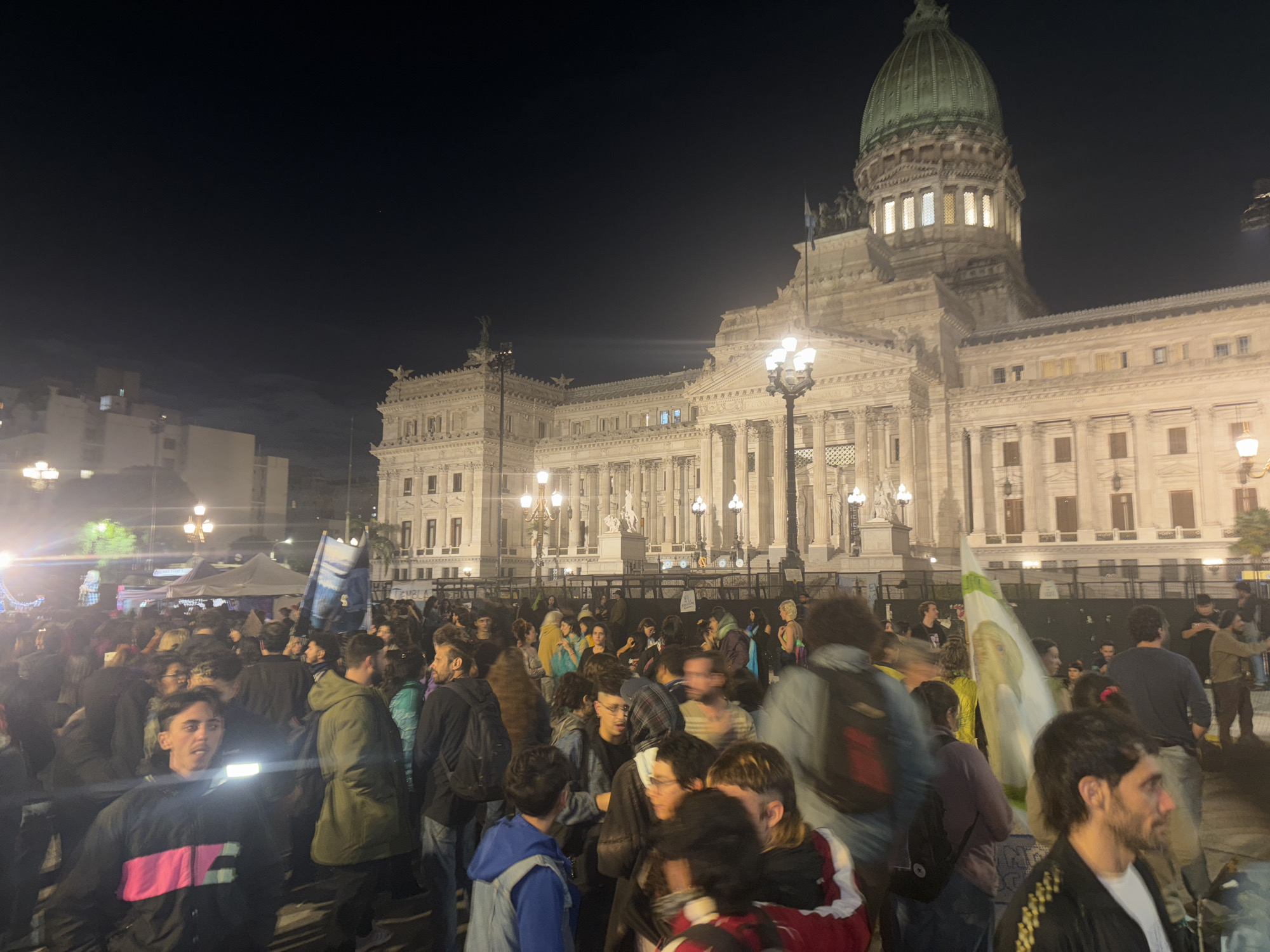 A nighttime crowd gathers in front of the Palace of the Argentine National Congress in Buenos Aires, Argentina. The crowd consists of people of various ages and clothing styles. Some individuals hold flags. The Palace of the Argentine National Congress is brightly lit with a green dome. The sky is dark. Streetlights illuminate the scene.