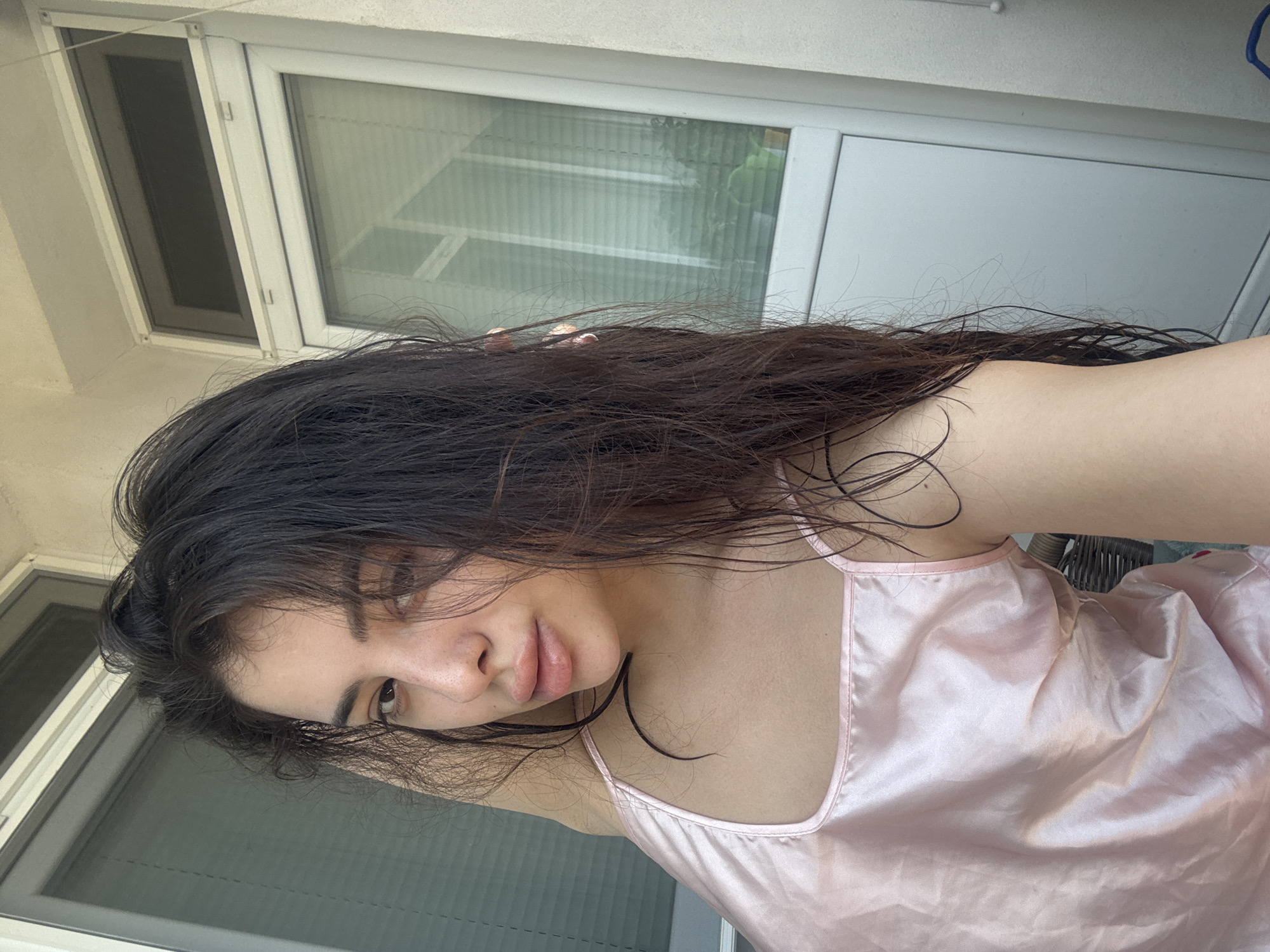 A young woman is depicted in a close-up, self-taken portrait, set indoors or on a balcony in Plovdiv, Bulgaria. She has long, dark brown hair, which appears somewhat wet or disheveled, falling over her shoulders and partially obscuring the left side of her face. Her complexion is fair, with visible freckles on her nose and upper lip. She is wearing a light pink, lustrous, possibly satin camisole or tank top. Her right arm is raised behind her head, with her hand positioned near her ear, likely holding the device used for the photograph. She looks directly into the camera with a neutral expression and slightly parted lips. The background features a white-framed window or glass door. Through the glass, reflections of interior vertical blinds are visible, and faintly, some greenery can be discerned outside, along with parts of an adjacent building structure. The scene is illuminated by natural daylight.
