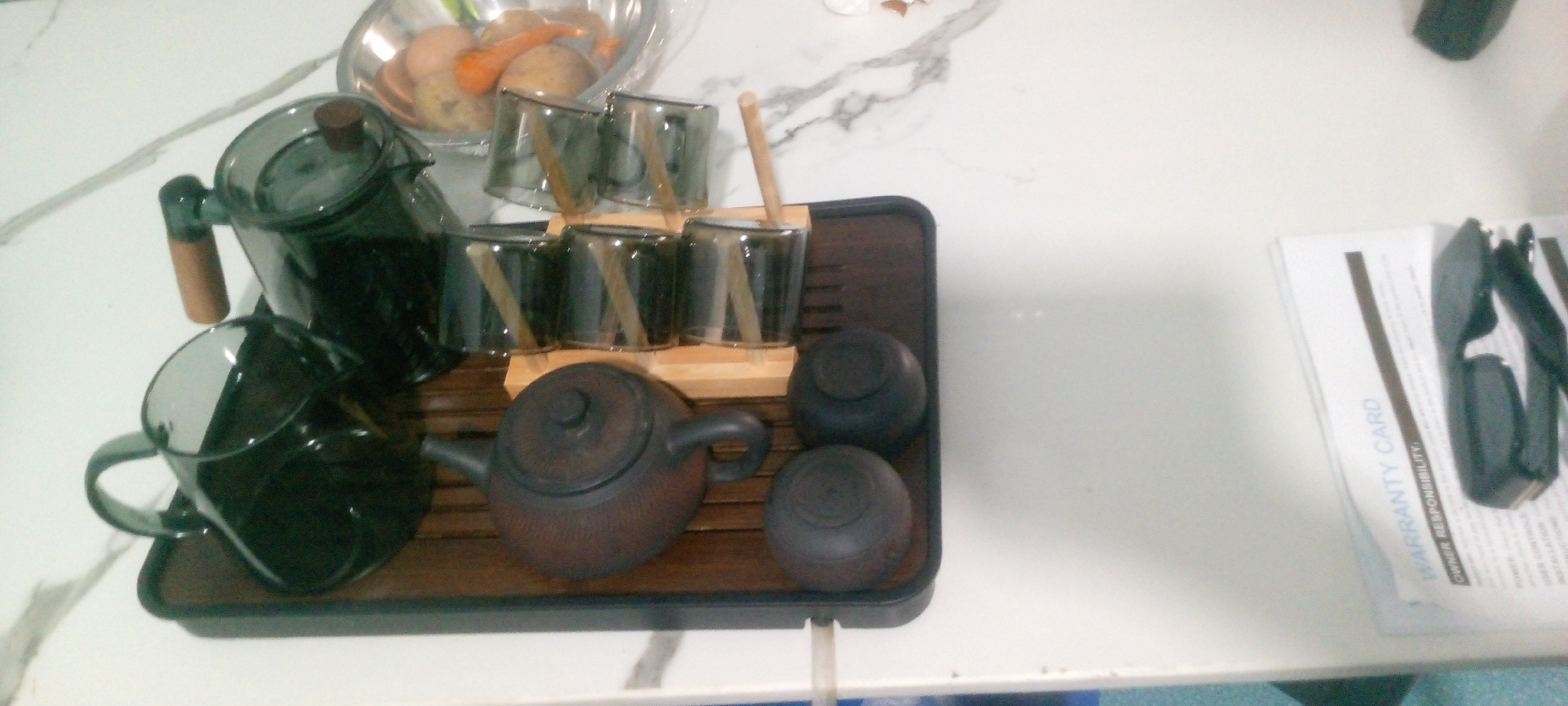 A tea set is displayed on a wooden tray on a white marbled countertop. The tea set includes a dark brown teapot, a clear glass teapot with a wooden handle, and six clear glass cups with wooden stirrers. Two small, round, dark-colored containers are also on the tray. In the background, a metal bowl containing potatoes and carrots is visible. To the right of the tea set, a white document with the words "WARRANTY CARD" is partially visible, with a pair of black sunglasses placed on top of it. The image appears to be taken indoors. There are no people in the image. The location of Indo, Nigeria is not deducible from the image.
