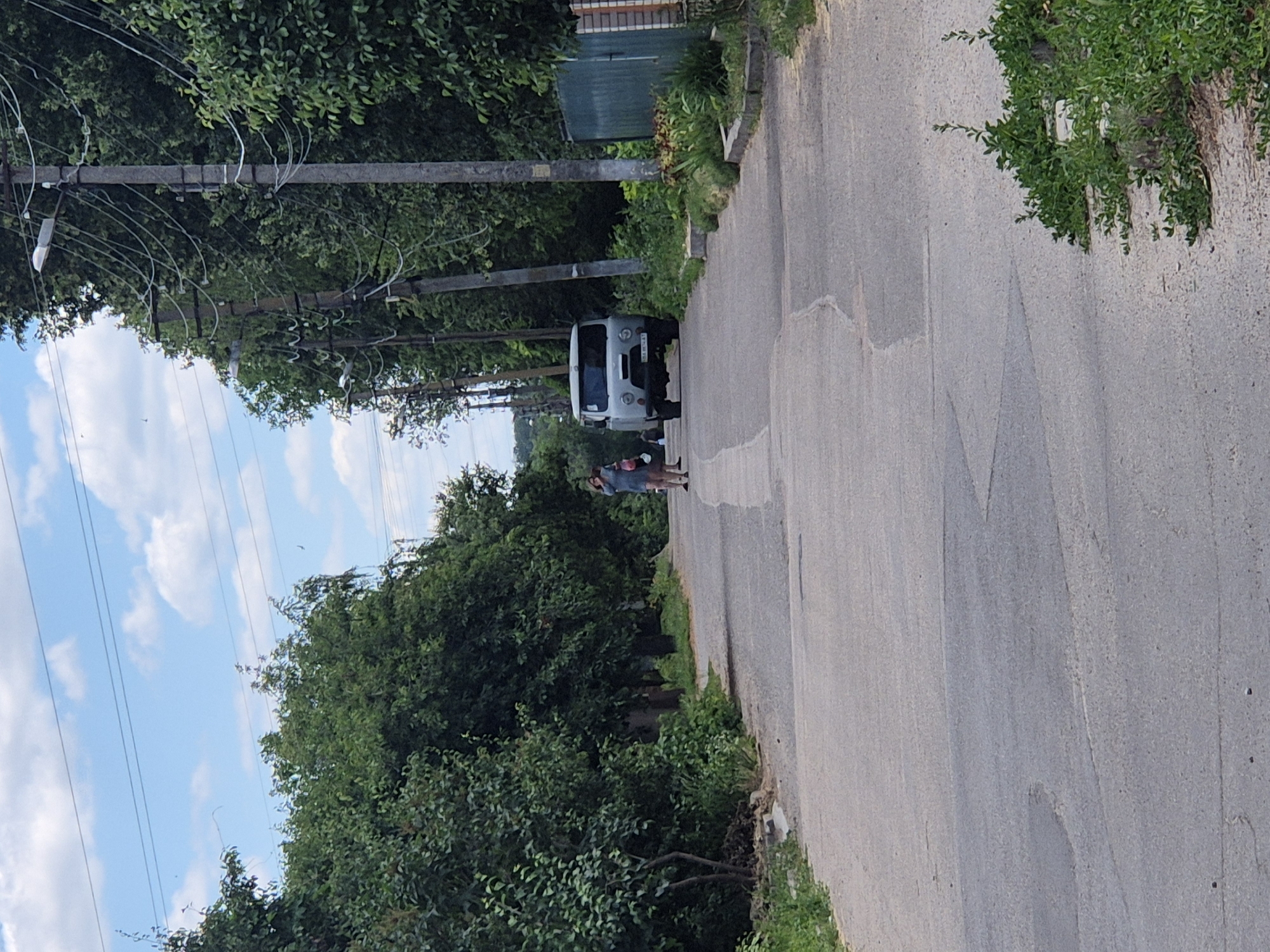 The image shows a rural road in Sozonivka, Ukraine, lined with trees and utility poles. A light-colored van is parked on the right side of the road, facing the camera. Two individuals are visible on the left side of the road, near the van. One person appears to be holding a bag. The road surface is asphalt and shows signs of wear. Greenery, including trees and grass, is abundant on both sides of the road. The sky is partly cloudy with a blue hue.