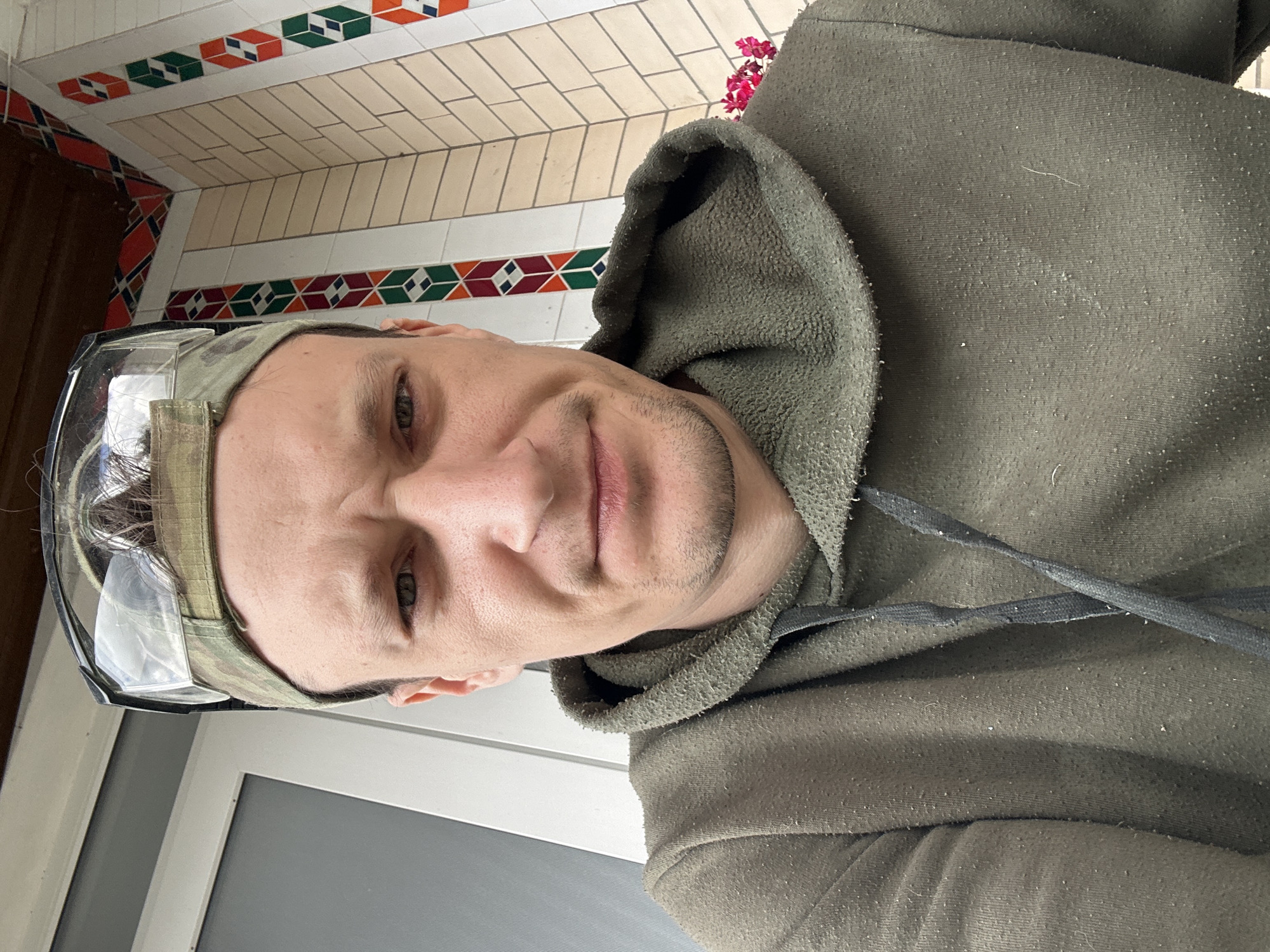 A close-up selfie shows a man wearing a green hoodie, a camouflage baseball cap, and clear safety glasses. He has a grimacing expression. The background includes a white door or wall on the left and a decorative tiled wall with red, green, and orange geometric patterns on the right. Some pink flowers are visible behind the man on the right side of the frame. The scene appears to be outdoors, possibly on a porch or near an entrance. The exact location within Makiv, Ukraine cannot be determined from the image alone.