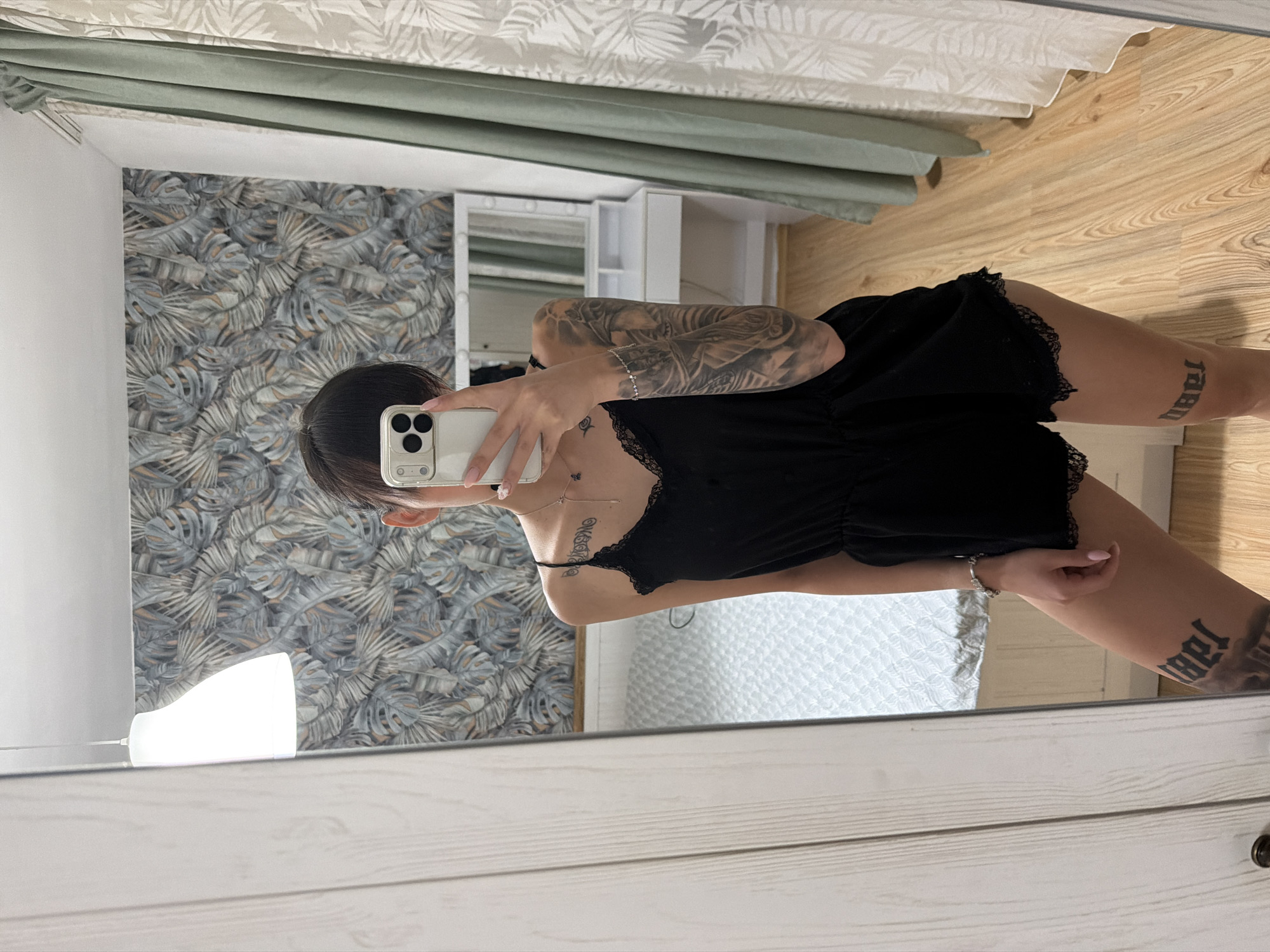 A person is standing in front of a full-length mirror in what appears to be a bedroom. The person is wearing a black romper with lace trim and has tattoos on their arm and thigh. They are holding a smartphone and taking a selfie. The room has a patterned wallpaper, a bed with a white quilted comforter, and curtains. A vanity with lights is visible in the reflection of the mirror. The floor is made of light-colored wood. The overall impression is a casual, personal snapshot taken in a private residence. The location is not specified within the image.
