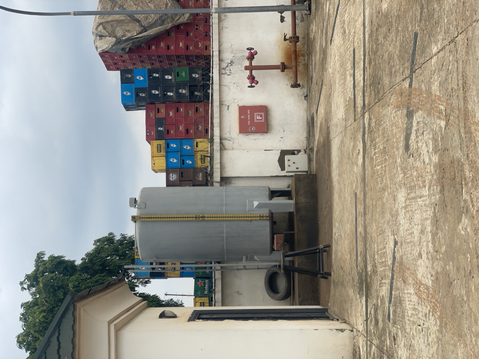 The image shows an outdoor scene, possibly a storage or industrial area, under an overcast sky, suggesting it might be daytime with cloudy weather. The primary subjects are stacks of various colored crates, a large cylindrical tank, a fire hose box, and a rusty pipe with valves. The crates, predominantly red, blue, and black, are stacked high and appear to be made of plastic, possibly for beverages. Some crates display text, including "AMSTEL" and "STAR".
A prominent feature is a large, gray, horizontal cylindrical tank, likely for storing liquid, with a yellow measurement scale on its side. Adjacent to the tank is a black tire resting on the ground. A red fire hose box is mounted on a white wall, featuring the letter "F" in a white circle. Above it, a network of rusty pipes with red valves is visible, a typical setup for fire suppression.
The setting appears to be in Abuja, Nigeria, as indicated by the location context. The ground is paved with rough concrete, showing signs of wear and tear, with several dark lines etched into its surface. In the background, lush green trees are visible, contrasting with the industrial elements. The overall impression is of a functional, utilitarian space, possibly a brewery's storage or operational area. No people are visible in the image, and the scene conveys a sense of stillness, with no clear activity taking place. The light is diffused, consistent with cloudy weather.