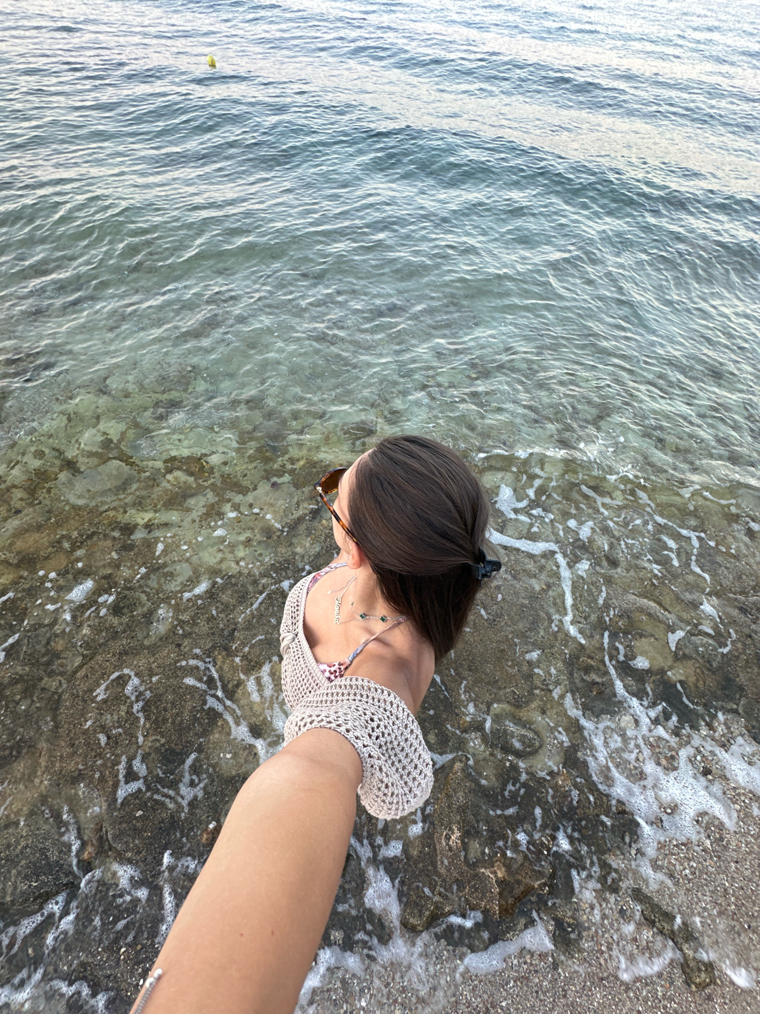 The image depicts a person standing at the edge of the sea, with their arm extended towards the camera, taking a selfie. The person is wearing a crochet top and sunglasses. The water is clear and reveals the rocky seabed. The scene appears to be a beach or coastline, likely in Kassandra Municipal Unit, Greece, given the context. The water is calm with small waves lapping at the shore.