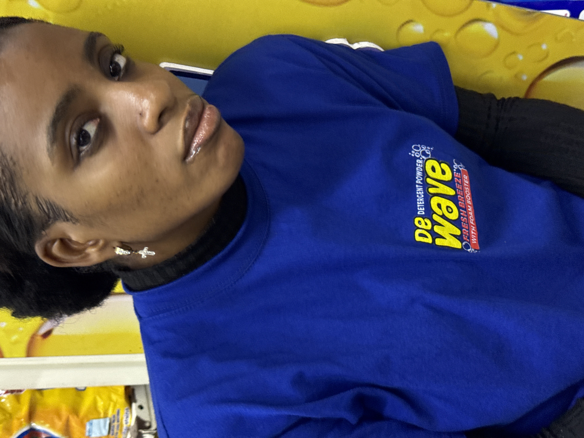 A young woman is indoors in Ado, Nigeria, wearing a blue t-shirt with a "de Wave" detergent powder logo on the front. She has a black top underneath. She has dark hair and is wearing cross earrings. The background includes a yellow surface.