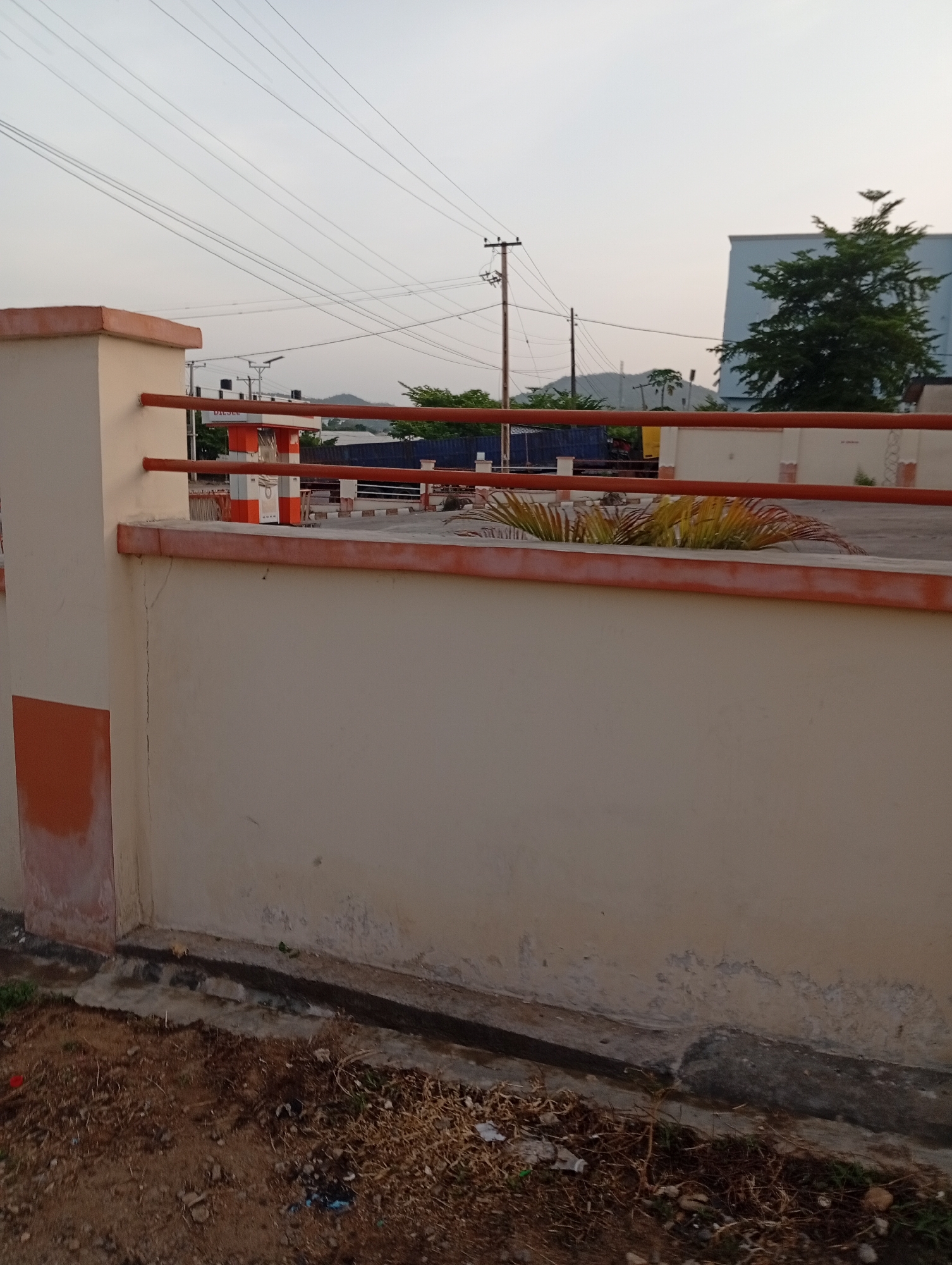 The image presents a low-angle view of an outdoor scene. In the foreground is a low concrete wall, painted light beige with a horizontal orange trim and vertical orange blocks on the sides. The wall has signs of weathering and discoloration, particularly near its base. Below the wall is an area of dirt and sparse vegetation. In the background, behind the wall, there is a paved area, visible through orange metal railings. In the far background, elements of the urban environment are discernible: utility poles with power lines, distant buildings, and trees. A blue building and vegetation can be seen behind the railing. A slight hill is visible in the distance under a hazy sky. The scene is bathed in soft, diffused light. Given the request that it's in Adankolo, Nigeria, no people are observed.