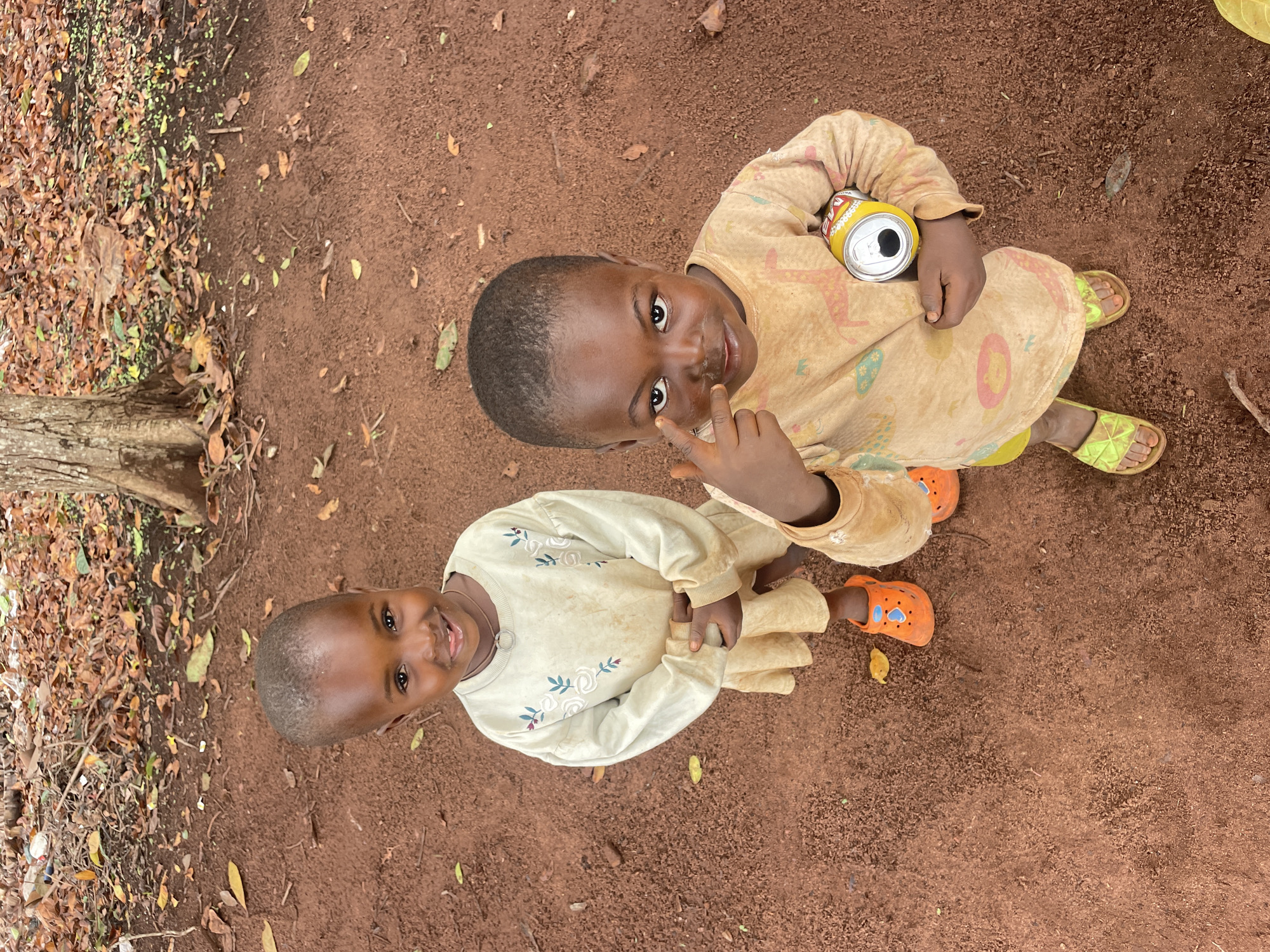 Two young children are standing on reddish-brown dirt near a tree and leaves. The child on the right wears a yellow patterned outfit and lime green sandals, holding an open can. They are pointing to their nose. The child on the left wears a light-colored long-sleeved outfit and orange clogs. Both children are smiling and looking upward. The location is Ogbe, Nigeria.