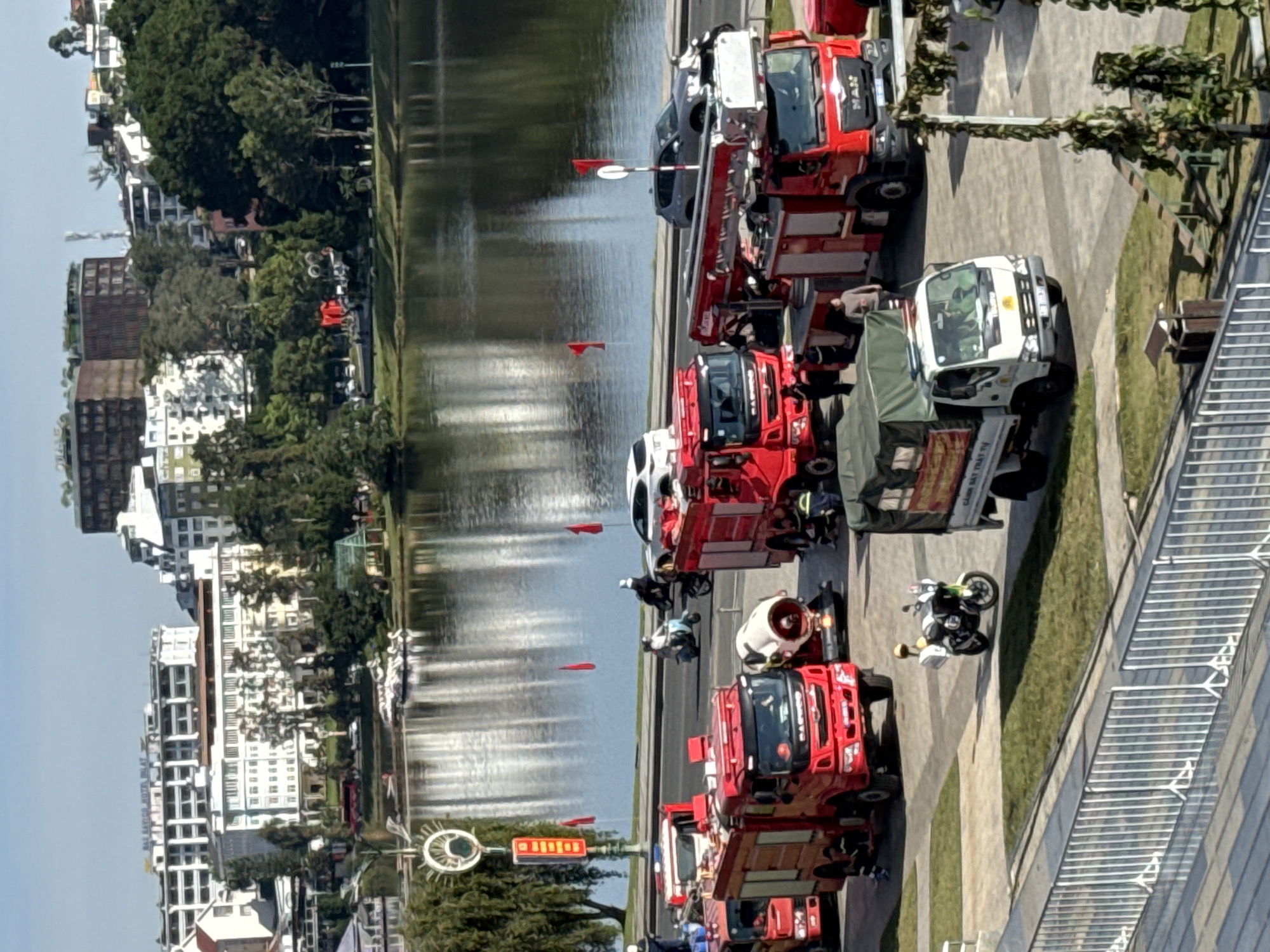 In the city of Da Lat, Vietnam, a fleet of red fire trucks and a white utility truck are parked along a waterfront, with a large lake reflecting the sky and surrounding buildings. Several individuals in uniform are visible near the fire trucks. In the background, a line of multi-story buildings, some with red roofs and others modern white structures, are nestled amongst lush green trees. The sky is clear and blue. The scene suggests a public event or a drill, possibly related to emergency services.