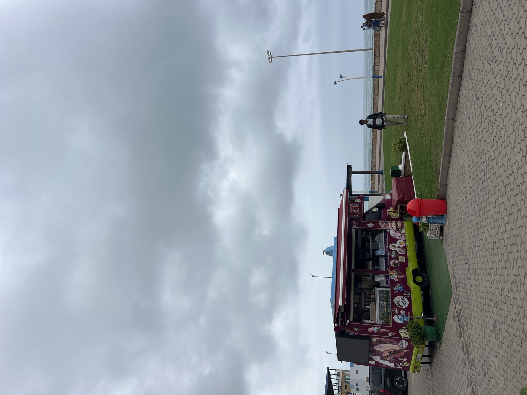 The image captures an overcast day at a coastal promenade in what is identified as M'Diq, Morocco. The primary subject is a vibrant food truck positioned on a paved pathway beside a strip of green grass. The truck features a dark red body with lime green lower accents and is extensively decorated with colorful cartoon illustrations of various food items, including ice cream, waffles, and possibly coffee. Text like "ICE CREAM" and "FASSIO" (or similar) is discernible on its side. A red trash can stands next to the truck, and a small patch of artificial green turf is laid out in front of it.
To the left of the food truck, beyond the grassy area, a person in a light top and dark jacket walks on the grass. Further in the distance, near the tranquil sea, a small group of people can be seen standing. The sky is largely grey and cloudy, indicating an overcast day with no direct sunlight. Tall, slender light poles are scattered along the promenade.
The setting is a public outdoor area, likely a walkway or park adjacent to the Mediterranean Sea. The paved path, made of interlocking grey and tan bricks, runs along the right side, bordering the well-maintained green lawn. In the background, the calm blue water of the sea stretches to the horizon, with faint outlines of land or structures visible beyond. The scene suggests a casual environment where visitors can enjoy refreshments by the sea.