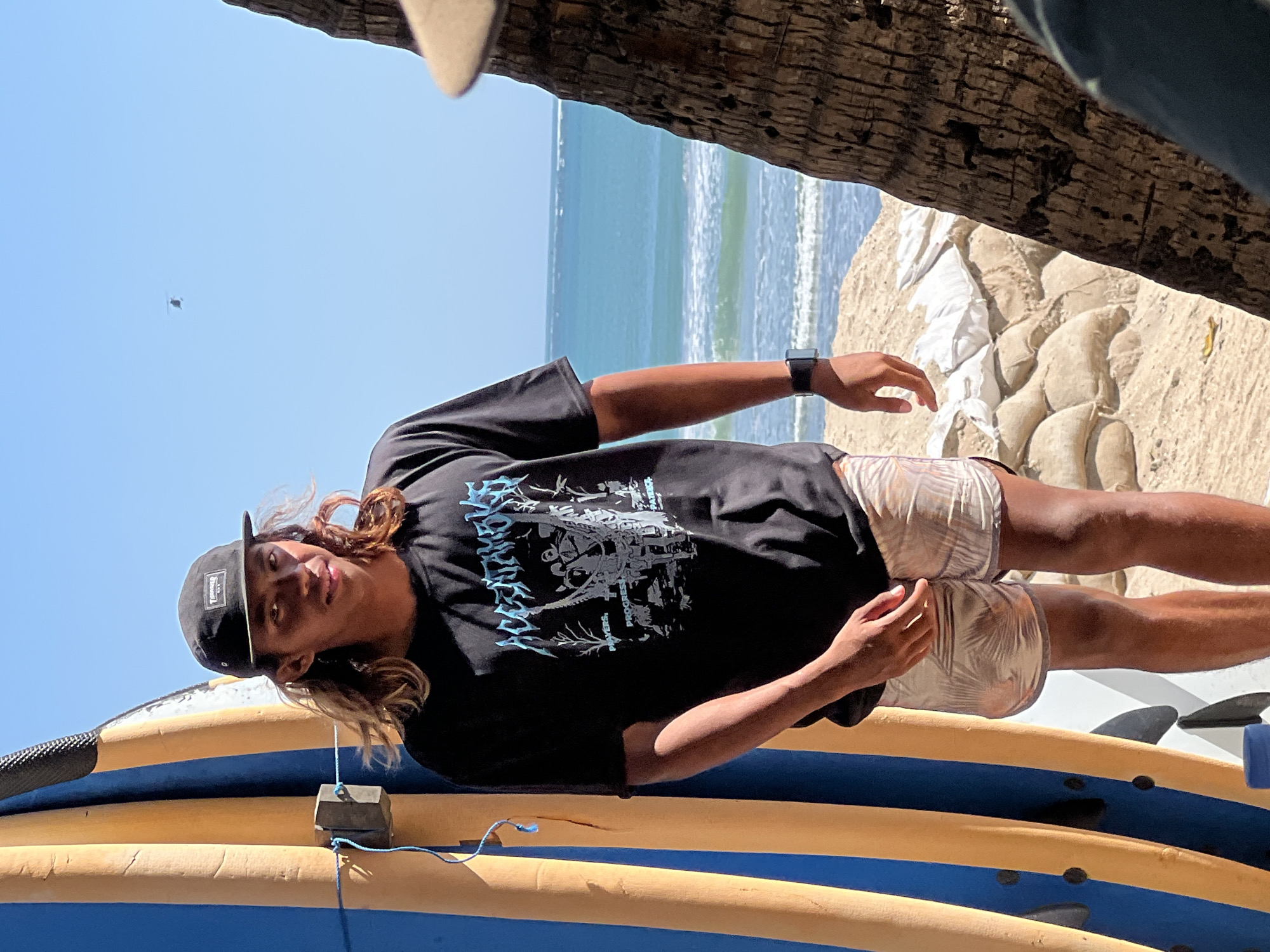 A young male with long brown hair and a black baseball cap stands on a sandy beach, leaning against a blue and yellow surfboard. He wears a black t-shirt with a graphic print and patterned shorts. The ocean with gentle waves is visible in the background, under a clear blue sky. Sandbags are piled up behind him, and a palm tree trunk is partially in the foreground on the right. This scene is located in Kuta, Indonesia.