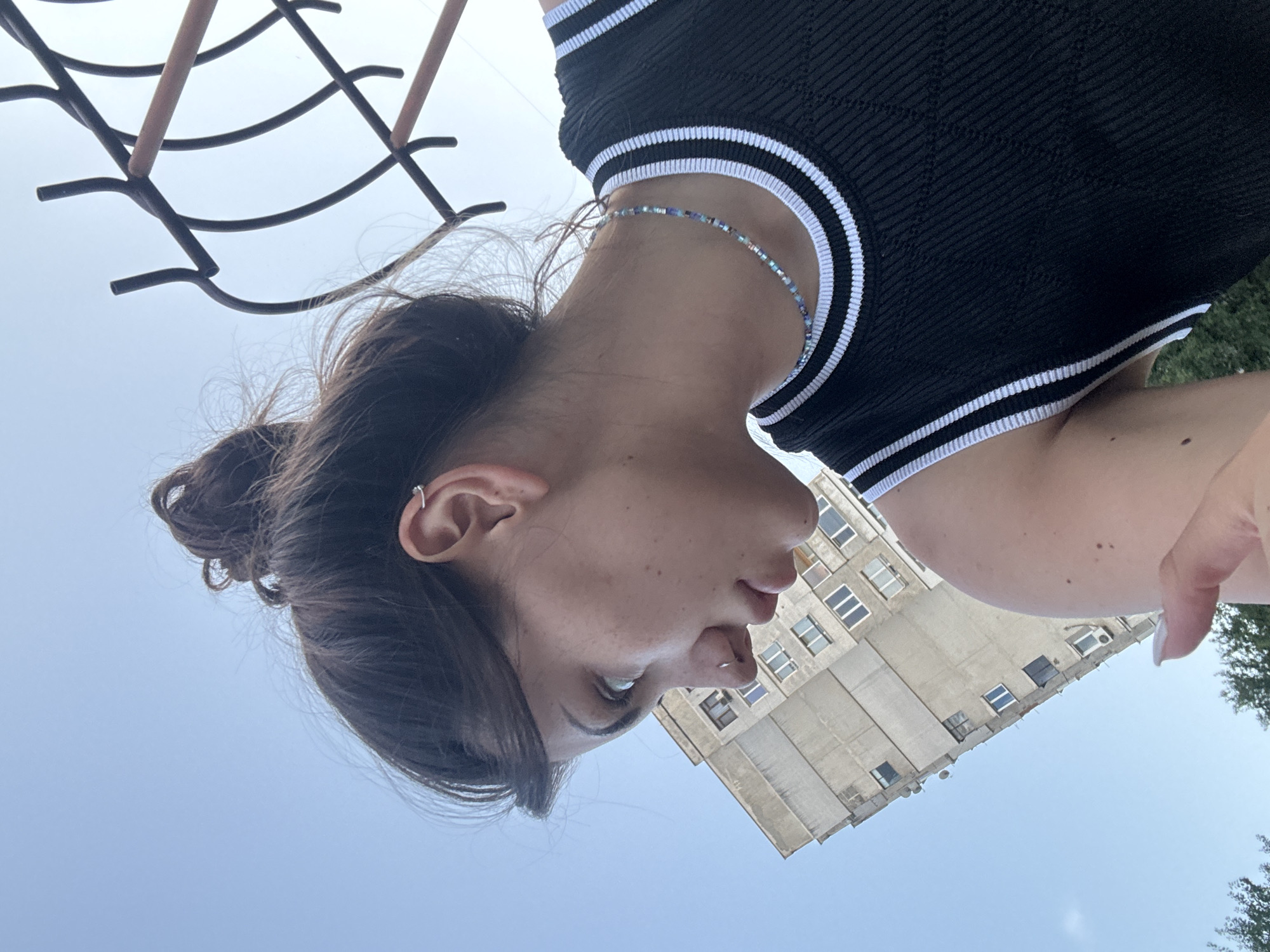 A young woman is captured in a low-angle shot, looking down and to her left. She has her hair pulled up in a messy bun and is wearing a black sleeveless top with white trim. A beaded necklace adorns her neck. In the background, a tall, concrete apartment building with numerous windows is visible, characteristic of Soviet-era architecture often found in Sofia, Bulgaria. A metal fence structure is also present in the upper right portion of the frame, partially obscuring the clear blue sky.