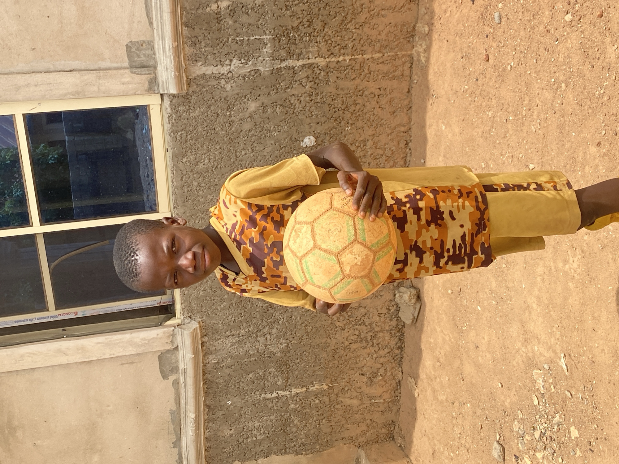 A young male individual stands facing forward in an outdoor, unpaved area in Rido, Nigeria. He has dark skin, short dark hair, and wears a yellow short-sleeved shirt under an orange and brown camouflage-patterned sleeveless garment, paired with yellow shorts or trousers and yellow footwear. He holds a worn, light orange/yellow soccer ball with green hexagonal patterns in both hands, displaying partial "FIFA STAND..." text. Behind him is a light beige, textured building wall featuring a horizontal molding and a coarser lower section. A window with a light-colored frame, showing "RONGTAI Global aluminum P-profile..." text on a mullion, is visible to the individual's right. The ground consists of light brown dirt with scattered small rocks and debris. The scene is illuminated by bright daylight.
