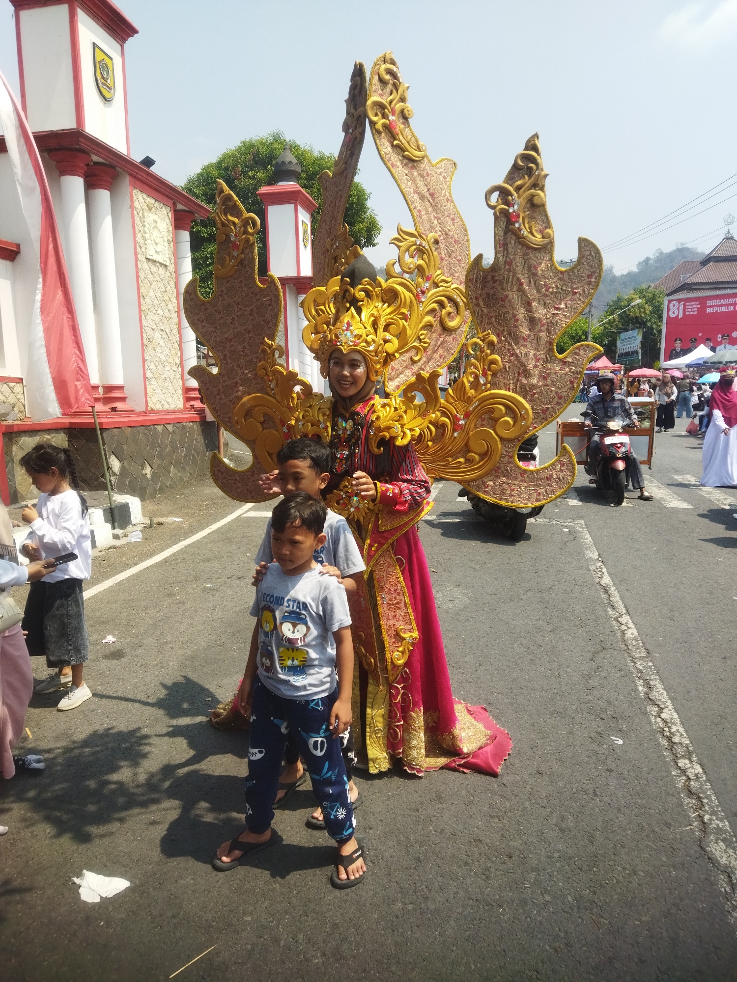 A woman in a decorative golden and red costume with a large, ornate headdress and wing-like adornments stands in the street. Two young boys, one in a gray t-shirt with cartoon characters and the other in a gray t-shirt with the words "ECOND STAR", stand in front of her. A woman in a white shirt and dark skirt is to their left, and another person in a pink garment is partially visible on the far left. In the background, a building with red and white trim, columns, and an emblem is visible. A red and white flag flies on a pole. To the right, a motorbike with a cart attached is being ridden by a man in a plaid shirt. A banner with "81 DIRGAHAYU REPUBLIK IN" is visible in the background, suggesting a national celebration. People are gathered on the street, with tents and umbrellas set up, indicating an event or parade. The scene appears to be in a city, with the building and banner suggesting it might be a public space in Wonogiri, Indonesia. The road is paved and marked with white lines.