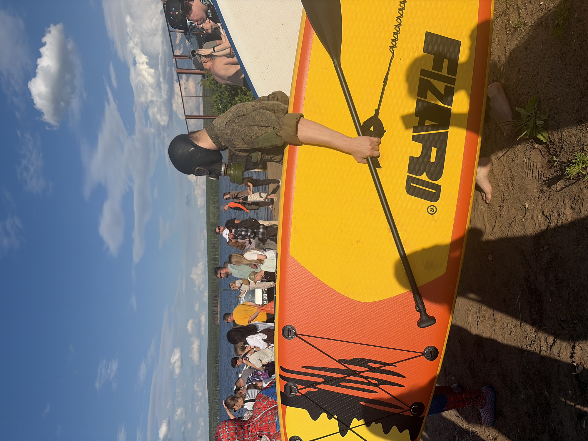 A group of people are gathered on the shore of a body of water, with a paddleboard in the foreground. A man in a military-style uniform and a black gas mask is holding the paddle, which is attached to the yellow and orange paddleboard. The paddleboard has the word "FIZARD" printed on it in large black letters. Behind the man with the paddleboard, a line of people are standing, some appearing to be on a boat or dock. One person in the background is wearing a Spider-Man costume. The scene appears to be outdoors on a sunny day, with blue skies and fluffy white clouds visible. The ground is sandy, with some vegetation growing nearby. The body of water is calm, with a treeline visible in the distance. The overall atmosphere seems casual and recreational. The presence of the gas mask and military uniform suggests a possible costume or theme for the gathering.