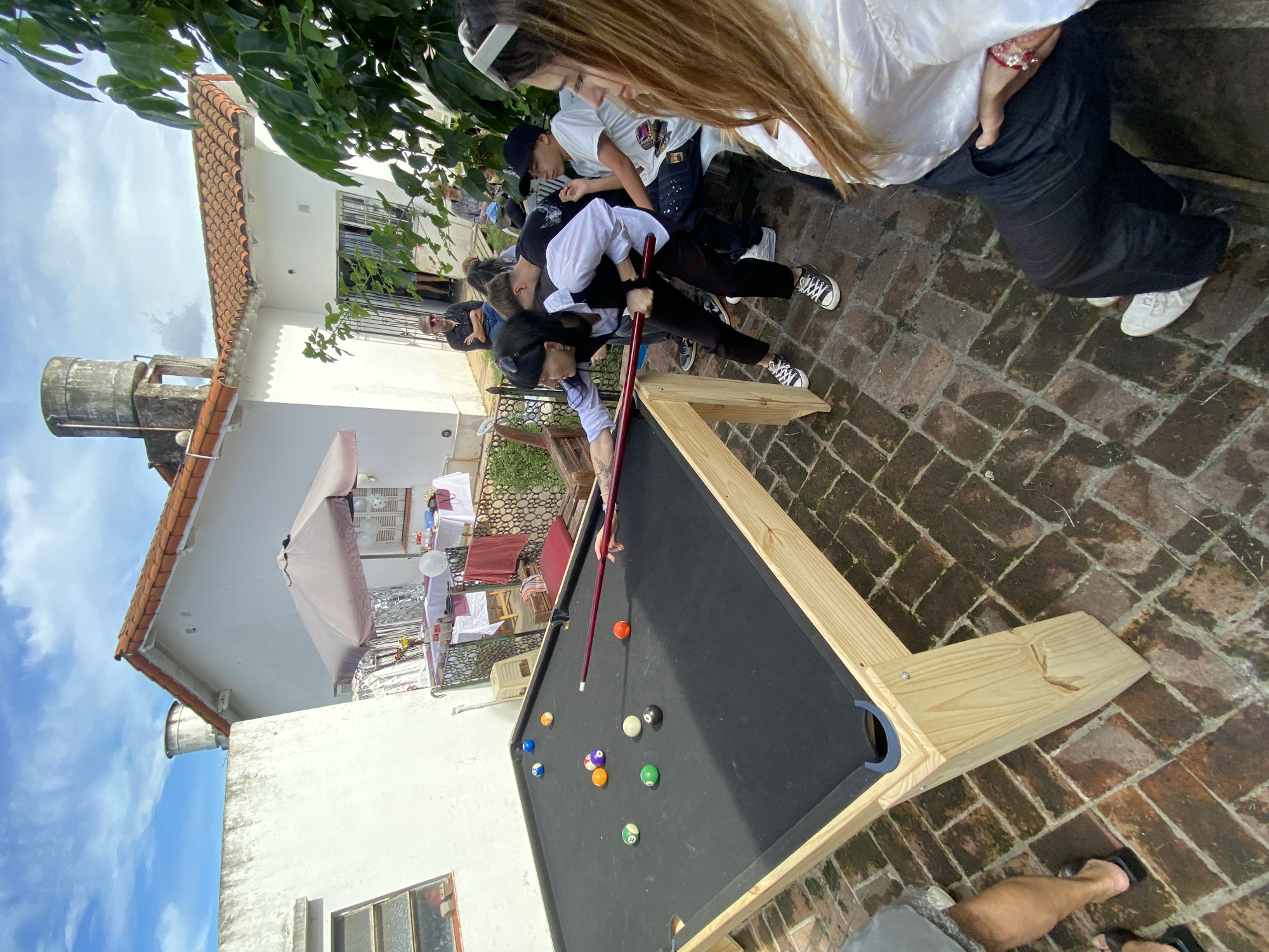 A medium, angled shot captures a gathering around a pool table in what appears to be an outdoor space, potentially a patio or backyard in Monte Grande, Argentina. Several individuals surround the pool table, watching someone take a shot. The pool table is a light wood construction with a black felt surface and standard billiard balls positioned. The location features brick or stone paving. In the background, there are white-painted structures, possibly houses or buildings, with visible architectural details such as a water tower and a sunshade over outdoor seating. Laundry can be seen hanging out to dry on a line. The sky, visible in the top-left corner, is blue with some clouds. The overall atmosphere seems casual and social.