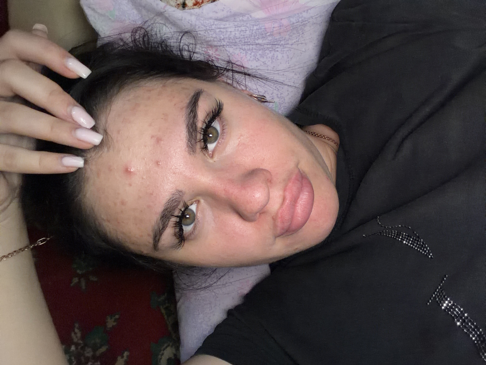 A close-up, high-angle selfie shows a young Caucasian woman lying down indoors, likely on a bed given the patterned fabric visible. She has dark hair, brown eyes, and prominent false eyelashes. Her skin appears to have acne. She is wearing a black t-shirt adorned with a rhinestone design. Her lips are pouty and slightly parted, and she is touching her forehead with her right hand, which has long, light-colored acrylic nails. A thin gold chain is visible around her neck. The background includes a red floral patterned fabric and a light-colored, patterned pillow or blanket. The image appears to be taken in a domestic setting in the city of Bolshebukovo, Russia.