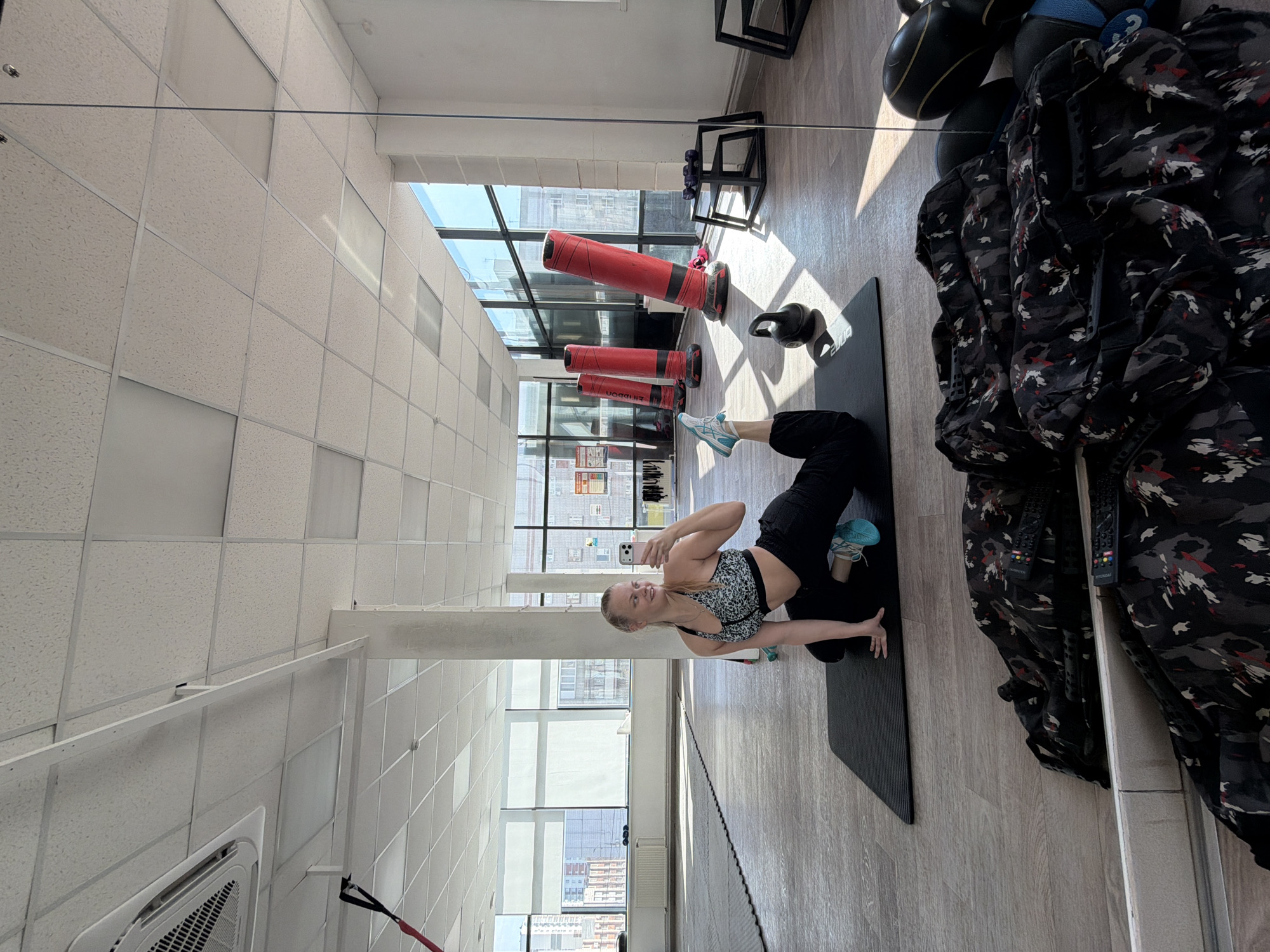 The image appears to be a selfie taken by a woman in what seems to be a gym or fitness studio. The gym has large windows showing a city landscape, potentially Izhevsk, Russia. The woman is dressed in athletic wear consisting of a sports bra and leggings. She is posing in a stretch or yoga position on a black mat. The gym environment is evident, with workout equipment such as punching bags, a kettlebell, and other items in the background. The image is taken indoors, during daylight hours, and the lighting is predominantly natural from the windows. The woman is holding a smartphone, presumably to take the photo. The flooring is light-colored wood. There's an apparent reflection from a mirror or glass surface on the right side of the image, indicating the presence of a reflective surface.