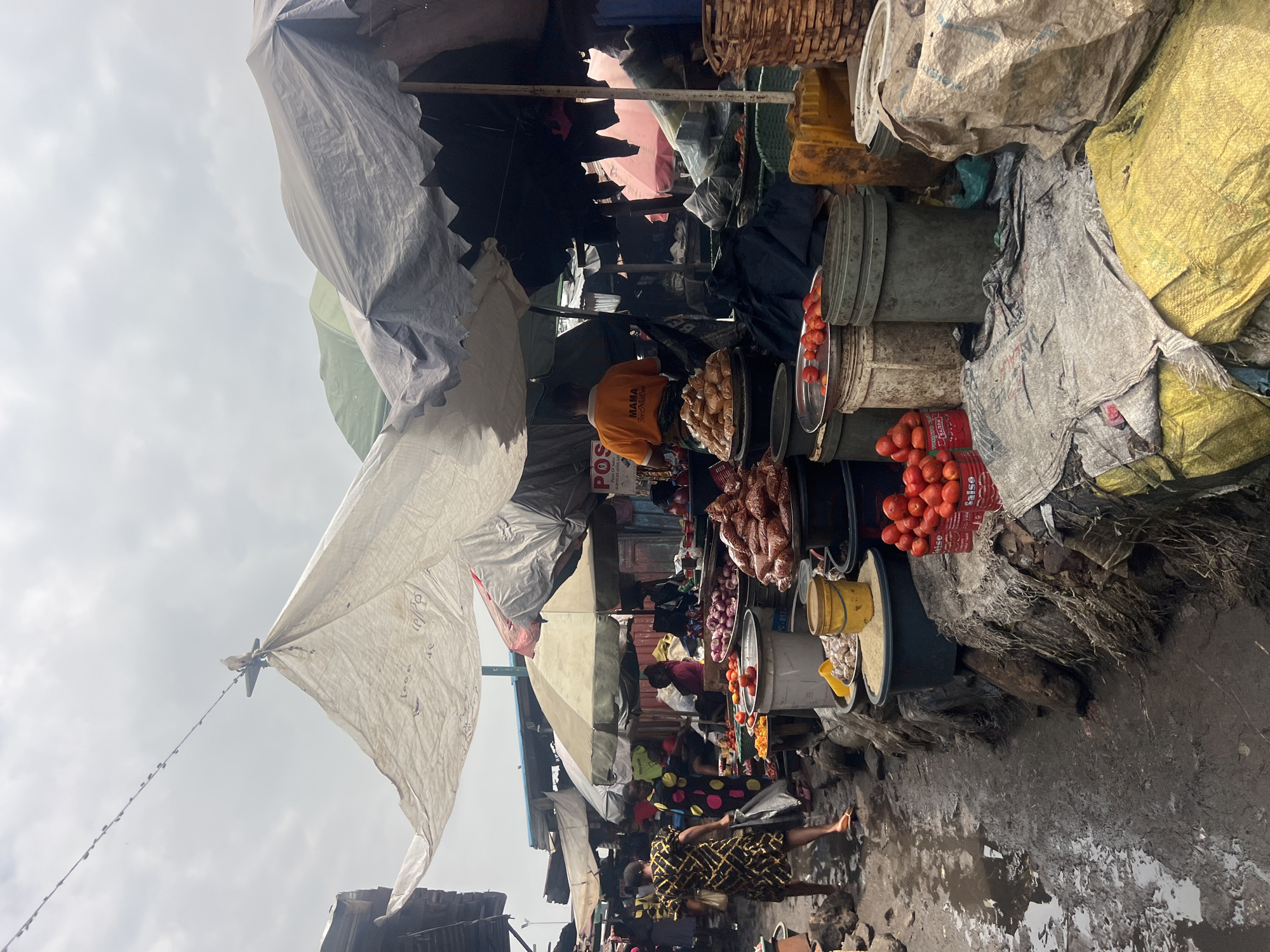 The image depicts a bustling outdoor market scene in Nkpor, Nigeria. The market appears to be a food market, with various fresh produce and goods displayed on stalls and the ground. Several people are visible, engaged in activities such as shopping, selling, and moving goods.
In the foreground, a woman in a patterned dress walks away from the camera, carrying a shopping bag and a purse. To her left, another woman in a polka-dot dress is interacting with a vendor. Numerous stalls are set up, covered by tarps and umbrellas to provide shade. These stalls display an abundance of fresh produce, including piles of tomatoes, onions, and other vegetables. Bags of what appear to be grains or dried goods are stacked on shelves and the ground.
The ground surface is wet and muddy, suggesting recent rain or inadequate drainage, which is common in many Nigerian markets. The overall environment is cluttered and appears to be a typical, lively, and somewhat unorganized market setting. In the background, more stalls and structures extend into the distance, with a mix of permanent and temporary shelters. The sky is overcast, contributing to the diffused lighting in the scene. The presence of large plastic containers and woven baskets suggests the storage and transportation of goods. A man in an orange shirt is visible behind a stall, possibly a vendor.