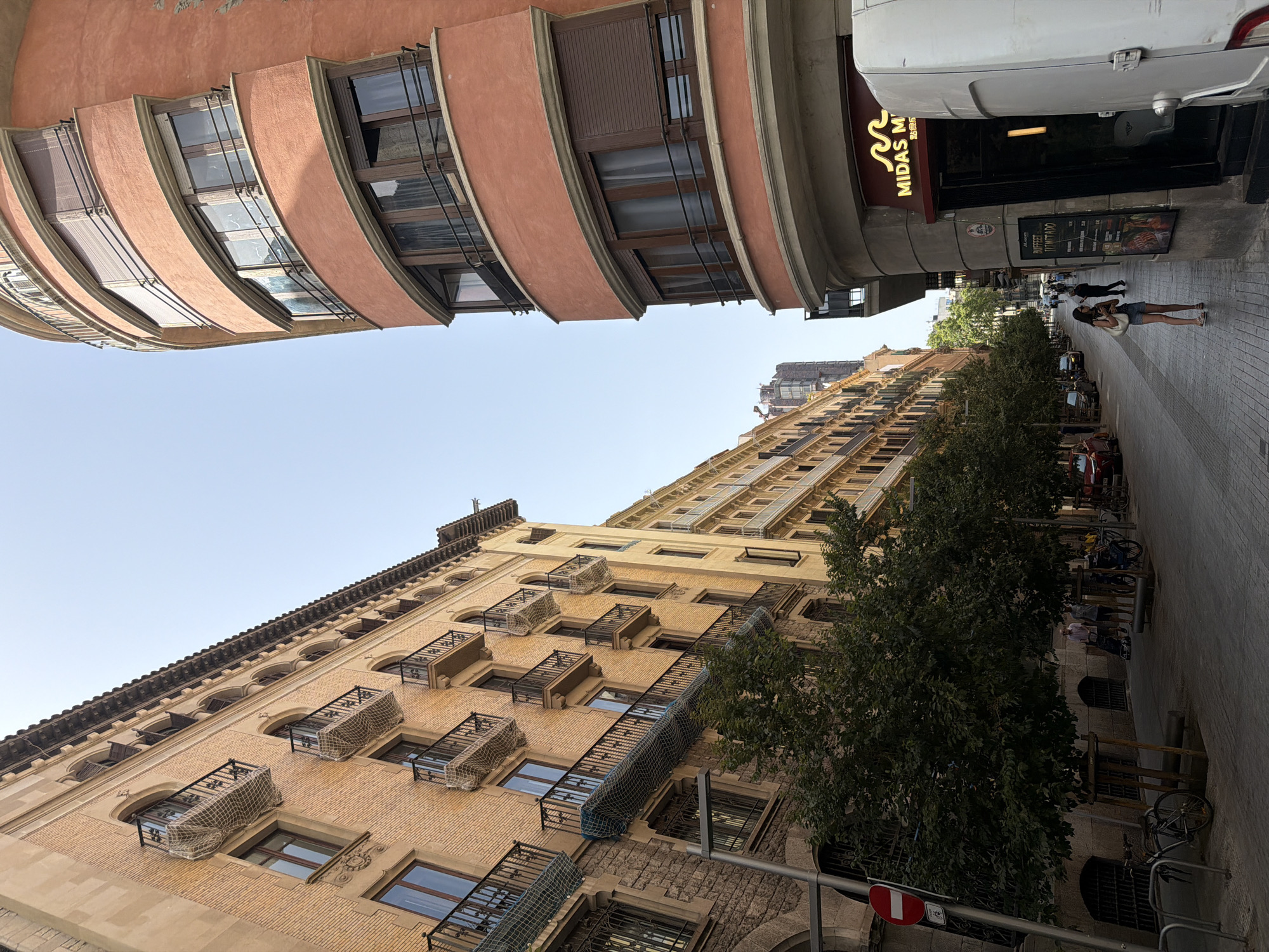 This is a street-level, upward-angle view of a city street in Barcelona, Spain, on a clear day. The street is lined with multi-story buildings, featuring a mix of architectural styles. On the left, a building with a light brown brick facade displays numerous balconies with black wrought-iron railings, some covered with netting. Adjacent to this, a building with a darker stone base shows similar features. On the right, a curved, salmon-colored building with rounded balconies and brown shutters dominates the foreground. A business sign, "MIDAS M", is visible above an entrance. The street itself is paved with cobblestones. Several trees with dark green foliage are planted along the sidewalk, providing shade. A few people are visible walking on the sidewalk, and parked cars line the street in the distance. A white van is parked in front of the business on the right. A bicycle is parked near the left side of the image.