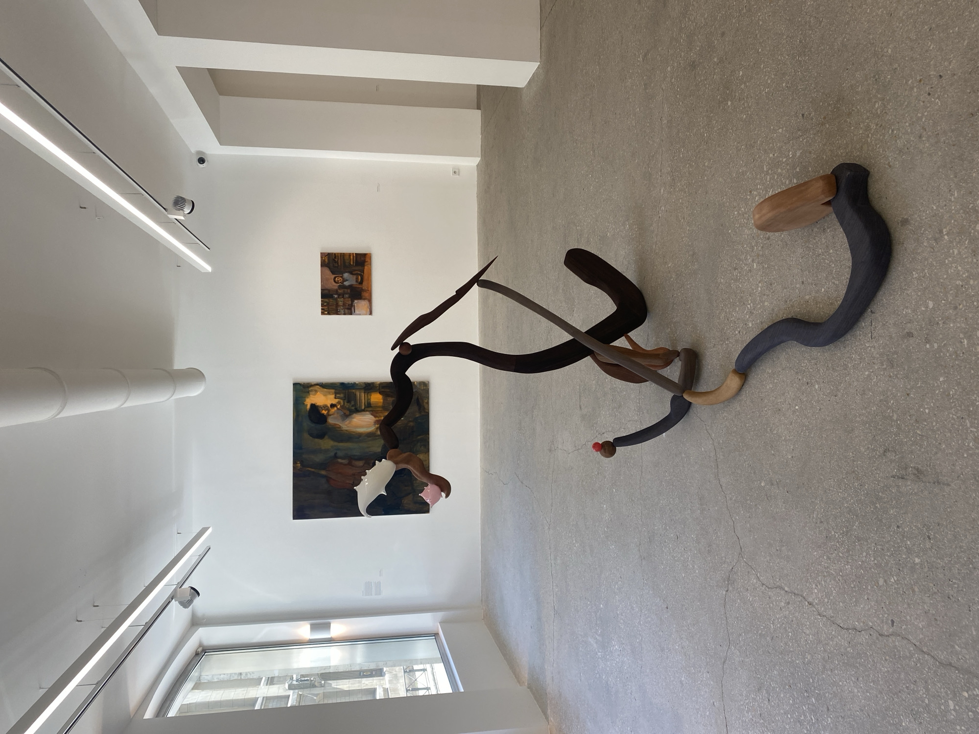 A high-angle shot shows the interior of a modern art gallery space. Two paintings hang on a white wall. The lower painting is a colorful, somewhat abstract depiction of figures, while the upper painting is a smaller, more abstract work in shades of brown. Dominating the foreground is a large, abstract sculpture made of various materials, including dark wood, light wood, and a grey-blue, snake-like element with a bulbous end painted red. The sculpture's elements intersect and twist, creating a dynamic and engaging form. The gallery's architecture is characterized by clean lines, white walls, and large windows. Long LED lights provide illumination. The floor is a polished concrete. The location is potentially in Paris, France, but there's no definitive element to confirm it.