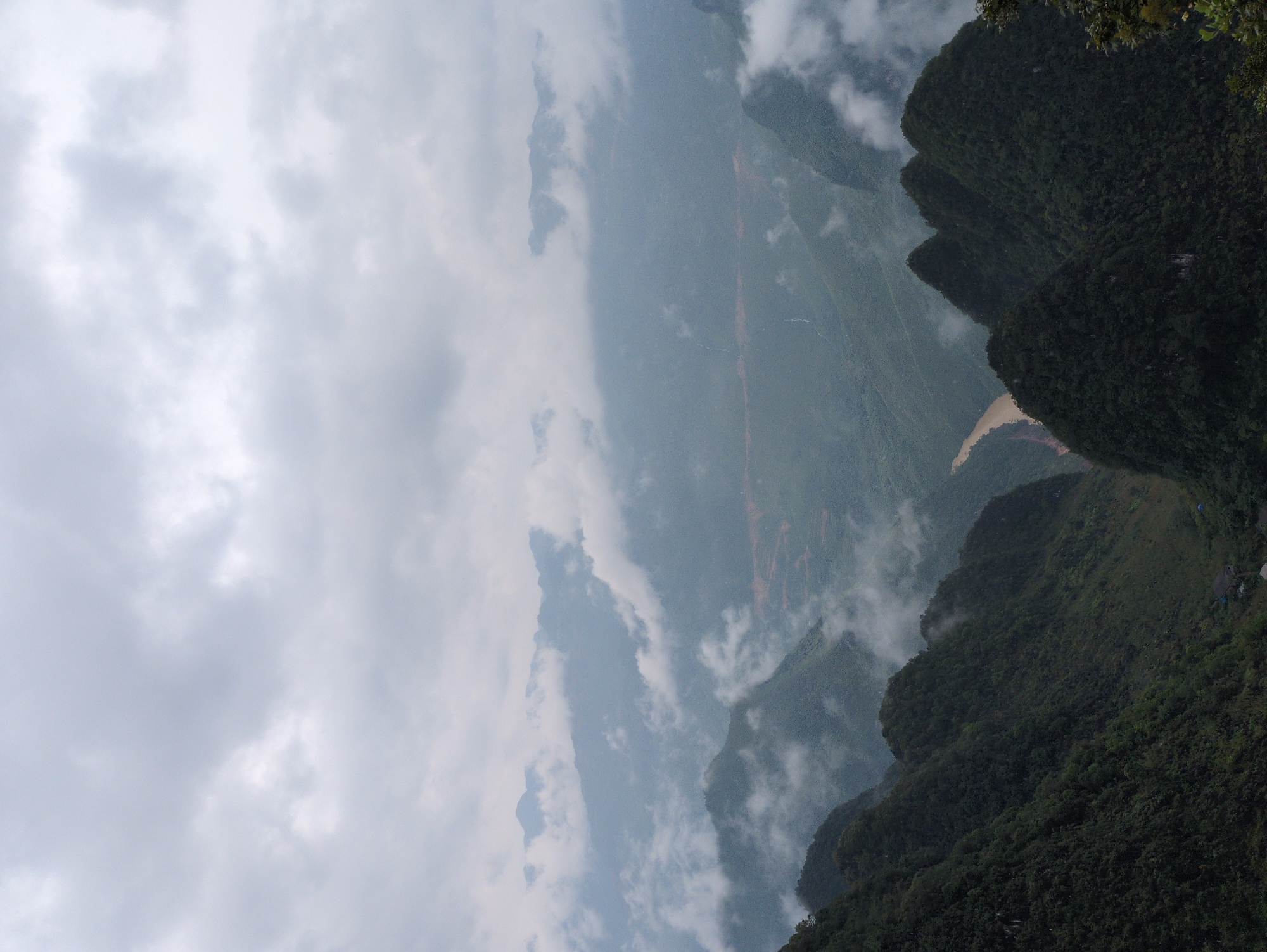 A high-angle, wide shot captures a dramatic, mist-shrouded mountain landscape in Đồng Văn Commune, Vietnam. The scene is dominated by dark green, densely forested mountains that rise steeply, their peaks obscured by soft, grey clouds and wisps of fog. A sinuous, muddy brown river snakes through a valley in the mid-ground, its banks lined with more lush vegetation. The rugged terrain is accentuated by the layers of mist that create a sense of depth and mystery. In the distance, the mountains recede into a hazy blue-grey expanse. The overall mood is serene and awe-inspiring, conveying the raw beauty and isolation of the Vietnamese highlands.