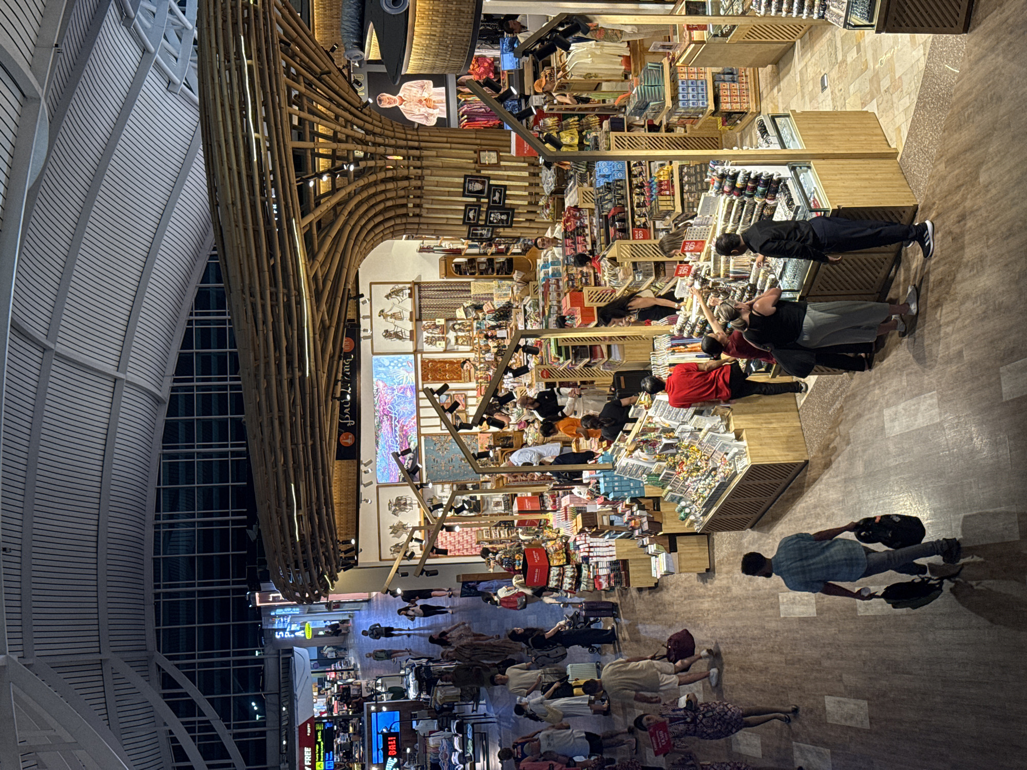 The image depicts a bustling interior scene within a duty-free shop, likely at an airport in Kuta, Indonesia, given the "Bali" signage visible on some stalls. The space is characterized by a large, arched bamboo structure serving as a prominent architectural feature. Numerous shoppers navigate through aisles filled with retail displays showcasing various merchandise, including clothing, souvenirs, and accessories. People are engaged in browsing, interacting with staff, and making purchases. The environment is well-lit with overhead lighting and spotlights illuminating the products. The flooring appears to be a polished tile or stone. The overall impression is one of a vibrant commercial hub catering to travelers.