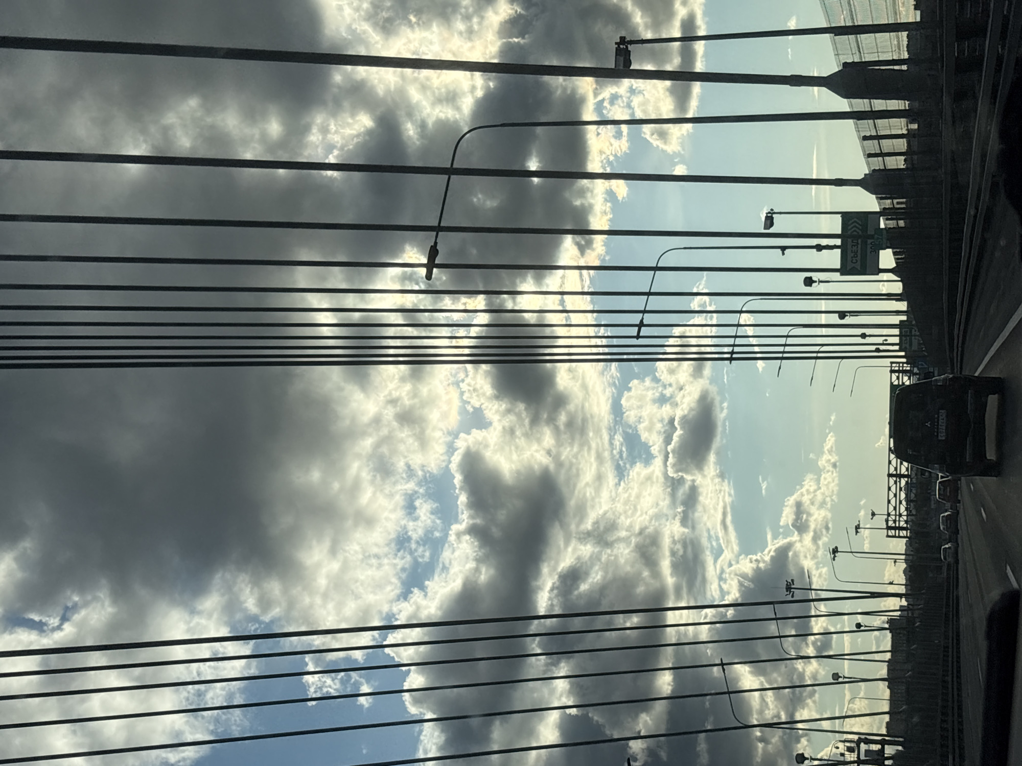 The image depicts a view from a vehicle traveling on a bridge, with the sky filled with dramatic clouds and sunlight breaking through. The bridge structure is characterized by numerous vertical cables that dominate the foreground, creating a strong geometric pattern against the sky. Numerous streetlights are visible along the side of the bridge. On the road, several vehicles are in motion, including a black SUV in the foreground. A green directional sign with Cyrillic text, indicating "Съезд" (Exit) and a route number, is visible on the right side of the bridge. The background shows the cityscape of Novosaratovka, Russia, with buildings and infrastructure visible in the distance under the cloudy sky. The overall scene suggests a dynamic, overcast day on a major thoroughfare within the city.
