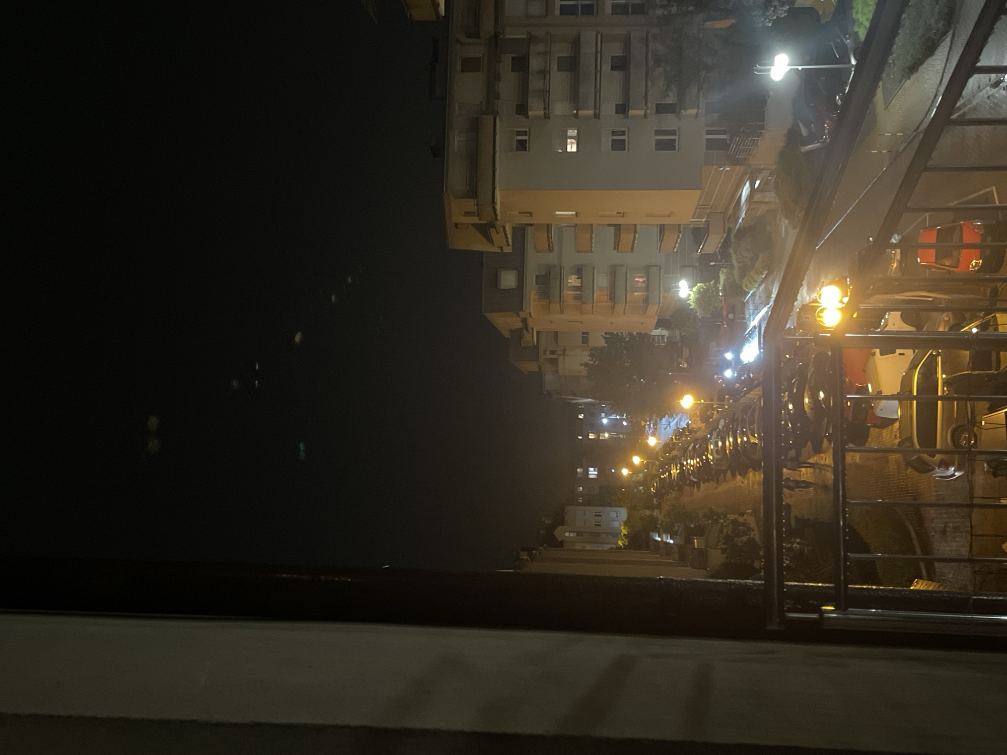 Captured at night, the image displays a city vista from an elevated perspective, potentially a bridge or high-rise. The architecture includes multi-story buildings with illuminated windows. A row of streetlights casts a glow along the roadway. Reflective surfaces hint at a waterfront presence, reflecting lights onto the water. In the foreground, a railing and a concrete structure suggest the viewpoint's edge. Numerous vehicles and motorcycles park alongside the street. The buildings seem to be in a grid layout.
