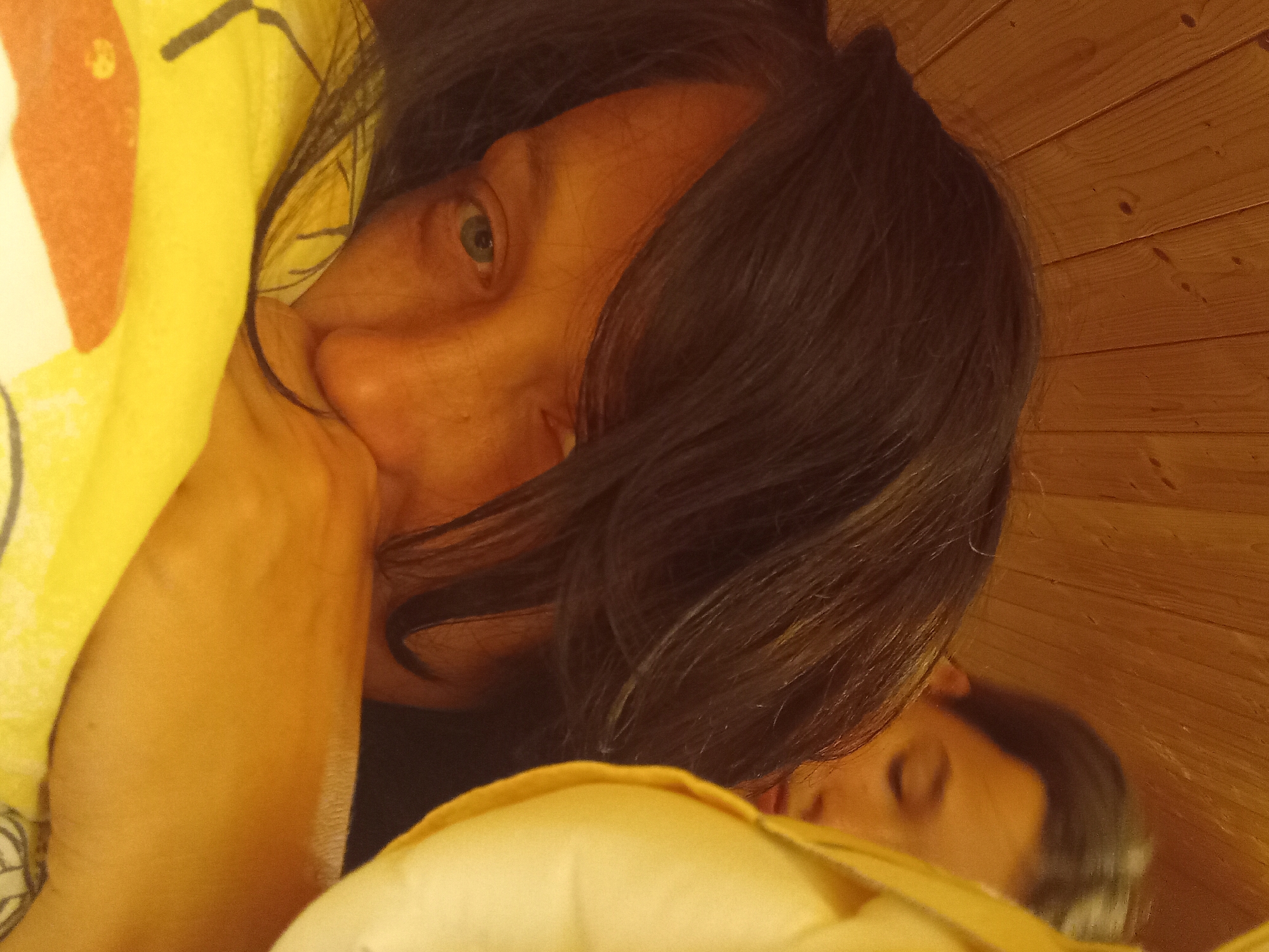 The image shows a close-up of a person's face, partially obscured by dark hair and a yellow blanket. The person is lying down, with their head resting on the blanket. Their eyes are open and looking towards the camera. A portion of another person's face is visible in the background, in reflection, suggesting the photo was taken in a mirror. The background is a wooden wall. The scene appears to be indoors. It is not possible to determine the location in Zuberec, Slovakia from the image.