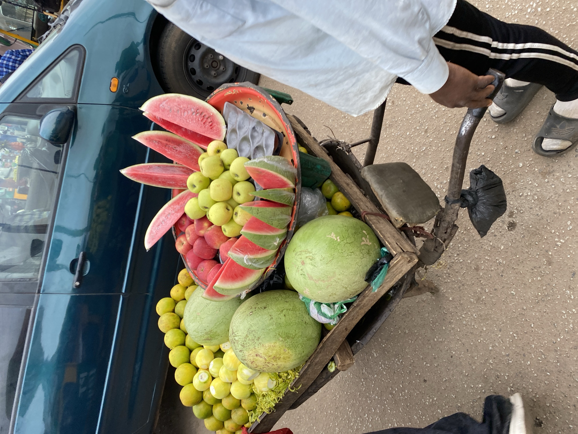 The image captures a street vendor in Bukuru, Nigeria, offering fruits and watermelons from a pushcart. The cart, constructed from wooden slats, holds a vibrant array of produce including whole watermelons, sliced watermelons, green apples, red apples and lemons.
The vendor, only partially visible, wears a white shirt and black pants with white stripes. The vendor's feet are clad in sandals. The cart is positioned near a dark green vehicle. The environment consists of paved street. The scene indicates a commonplace commercial interaction, typical of street vending in urban Nigeria.