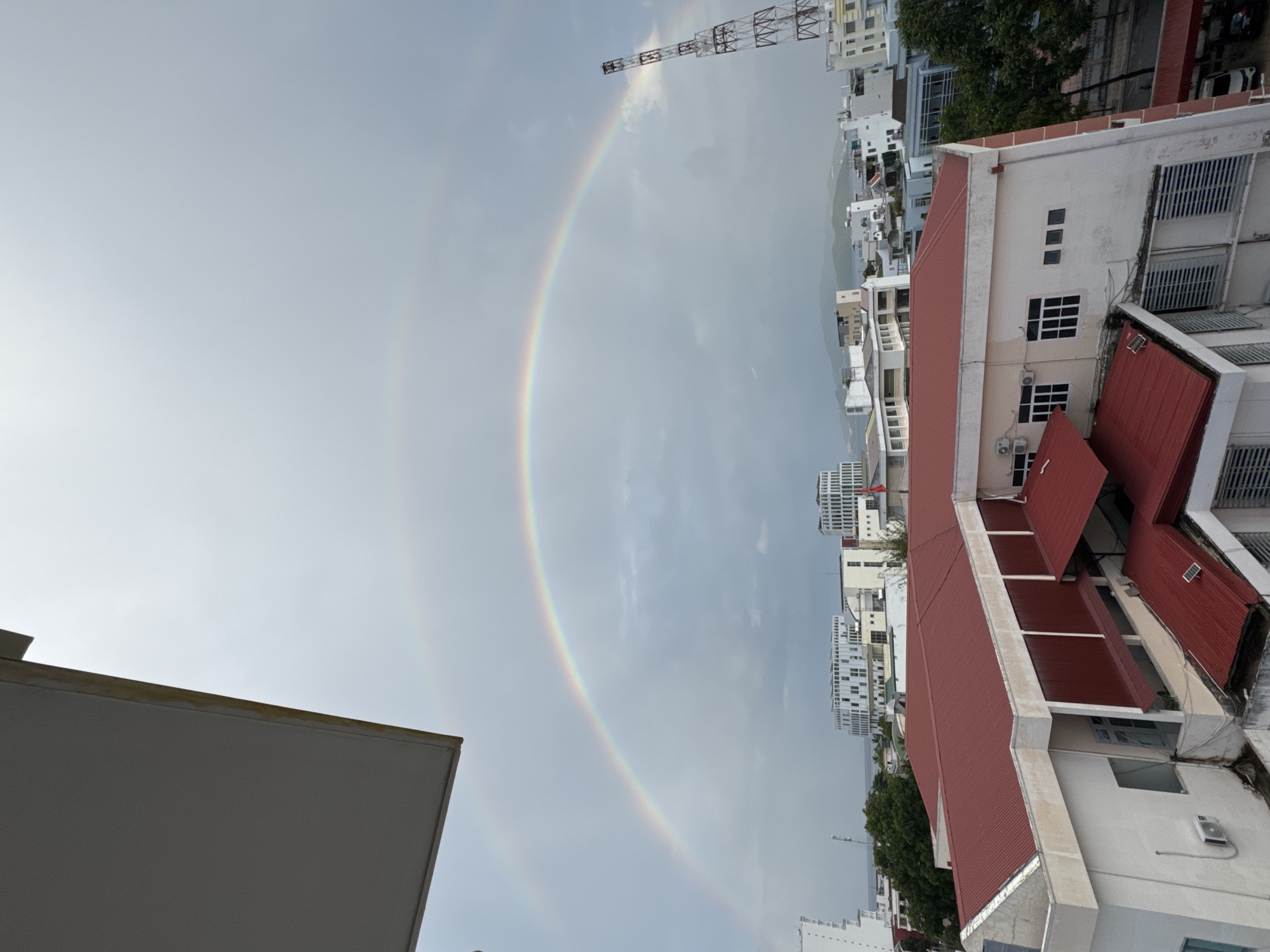 An overcast sky over the city of Nha Trang, Vietnam, is illuminated by a prominent double rainbow. The primary rainbow arches across the sky, its colors muted by the diffused light. A fainter, secondary rainbow is visible above it. Below the rainbow, a cityscape unfolds with various buildings, including a large structure with a red corrugated metal roof in the foreground. In the background, modern buildings with white facades and grid-like windows are visible, alongside a tall, lattice radio tower. The overall impression is one of a city under a dramatic sky following a rain shower.