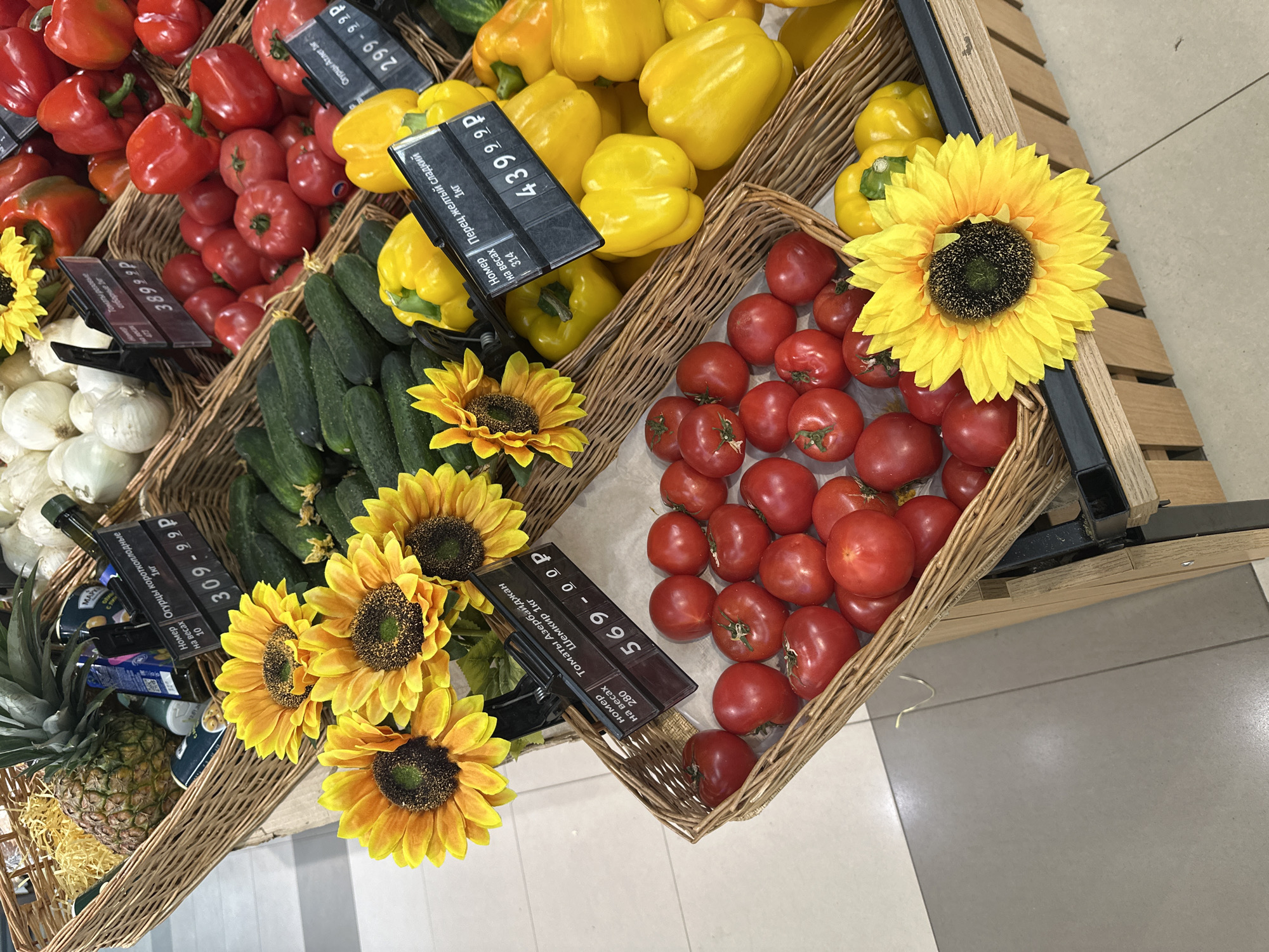 The image shows a display of produce in a grocery store. The items are arranged in wicker baskets. The products include red bell peppers, yellow bell peppers, tomatoes, cucumbers, onions, and pineapples. There are yellow sunflowers used as decorative elements in the baskets. Price tags are visible, displaying prices in Russian Rubles (e.g., 569-00 р). Based on the product names (e.g., "Томаты Азербайджан Шемкир 1кг") the store is located in Moscow, Russia. The floor is tiled in white and gray.