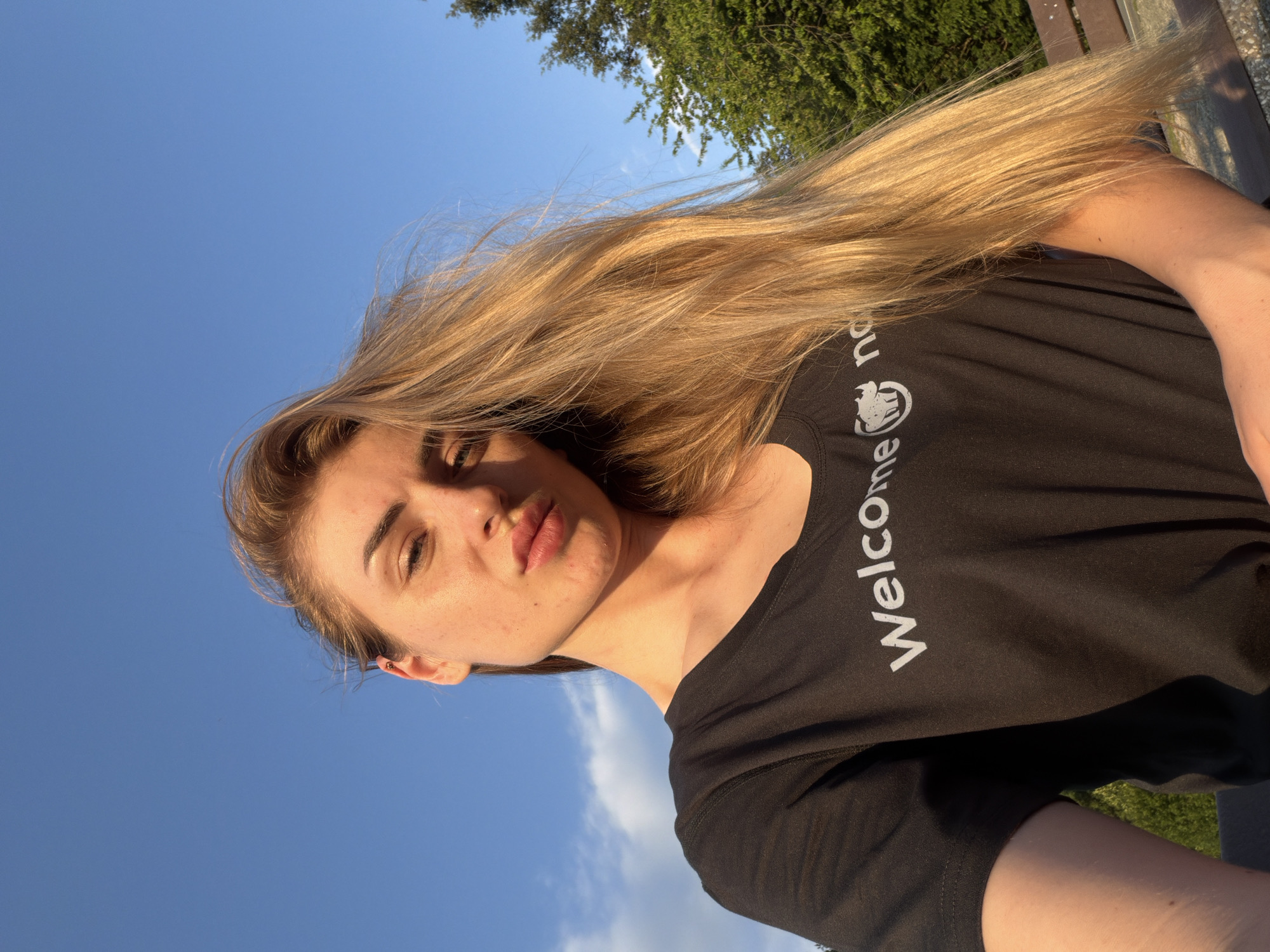 A medium shot photo shows a young woman with long, blonde hair posing outdoors under a clear blue sky with sparse clouds. She is wearing a black t-shirt with the word "Welcome" and a rhino logo printed in white. Her lips are slightly pursed as she looks at the camera. Sunlight illuminates her face and hair. In the background, green trees and a brown wooden bench are visible. The scene appears to be a park or natural area.
