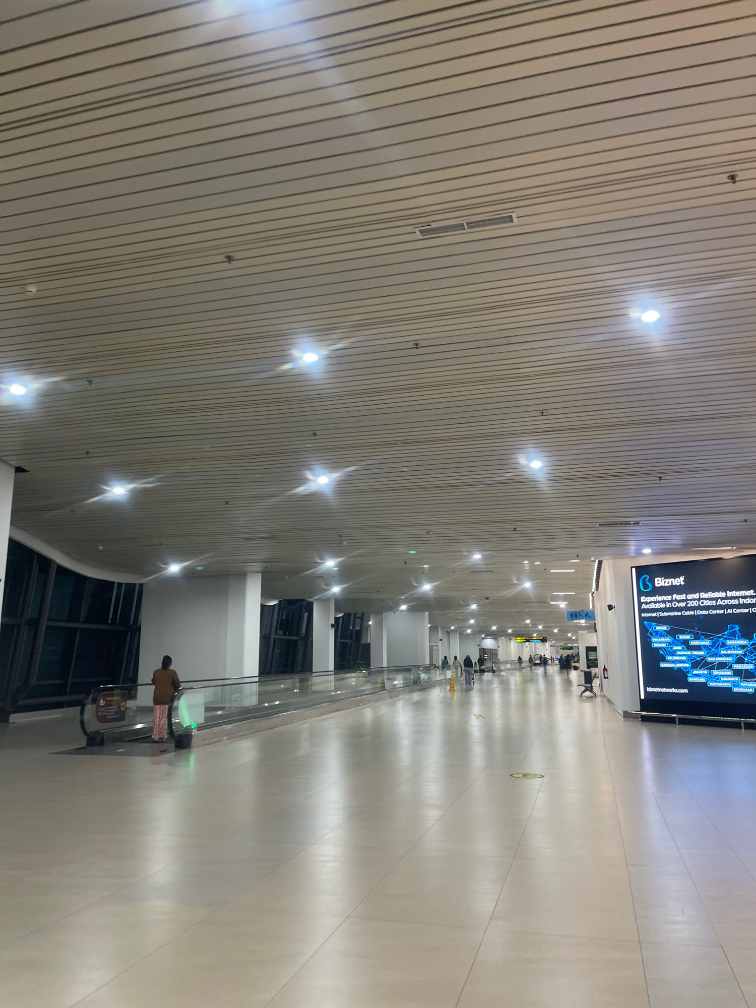 The image depicts the interior of an airport terminal, likely located in Rawa Burung, Indonesia, given the presence of "Biznet" advertising which is an Indonesian internet service provider. The scene is a long, brightly lit corridor with a polished, reflective floor. A moving walkway runs along the left side of the corridor, occupied by a person wearing a brown top and patterned pants. Further down the corridor, several other individuals are walking, and some appear to be standing. The ceiling is constructed with horizontal slatted panels, with numerous embedded lights and what appear to be air vents or sprinkler heads. On the right side of the corridor, a large digital display screen advertises "Biznet," showing a map of Indonesia and listing various cities. The overall environment suggests a modern, functional airport transit area.