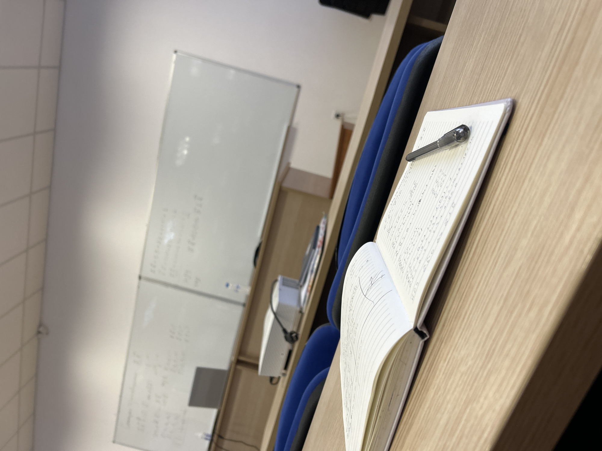 The image depicts an indoor classroom setting. There are rows of blue chairs arranged at desks. On a desk, there is an open notebook with handwritten notes and a pen resting on top. In the background, there is a whiteboard with writing on it and a projector. The specific location in Plovdiv, Bulgaria, is not determinable from the image.