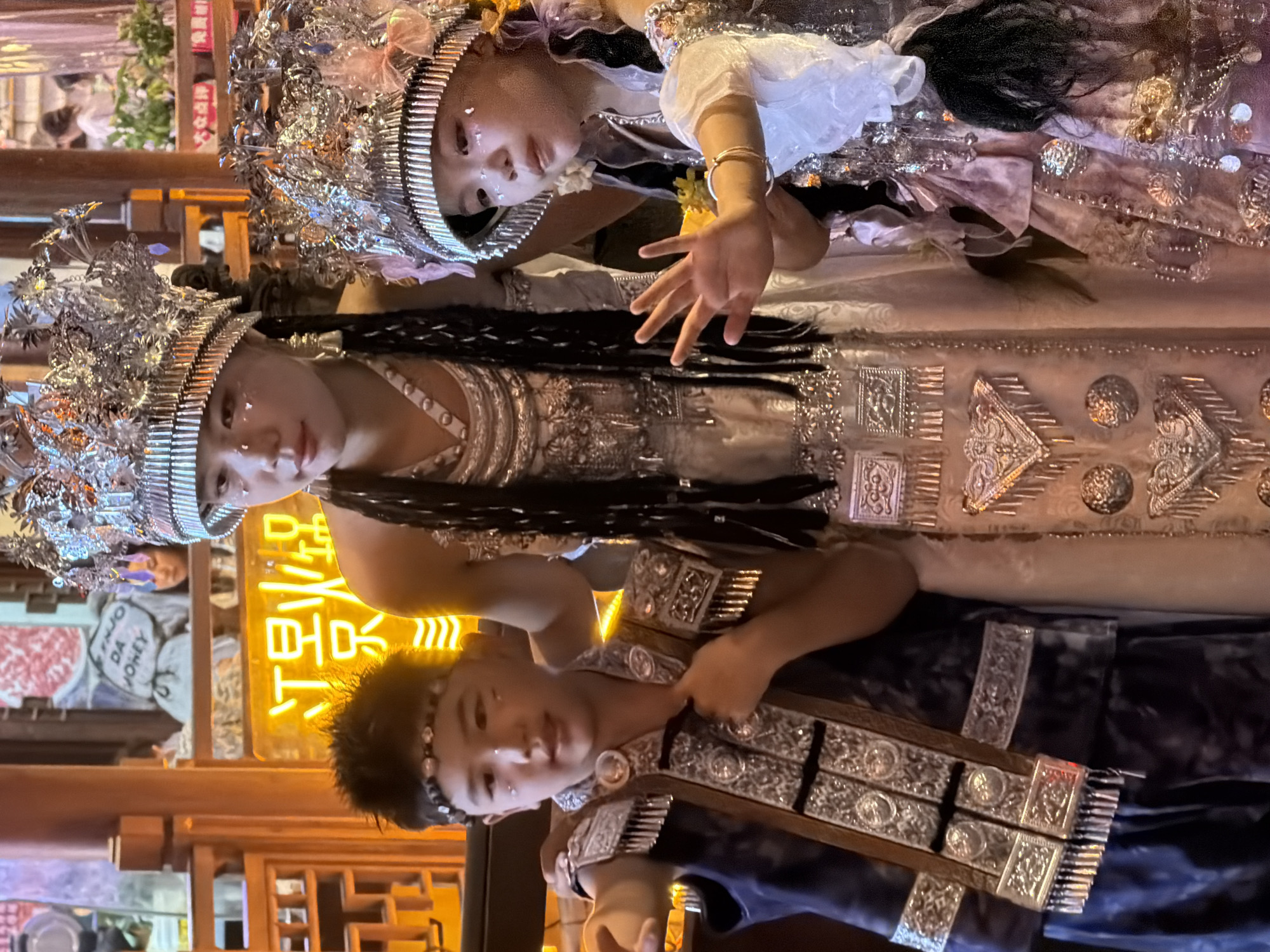 Three individuals, two children and one young adult, are posing for a photograph in an outdoor setting that appears to be a night market or entertainment venue in Yuzhong District, China. The individuals are adorned in elaborate, traditional-style costumes and headdresses with intricate silver ornamentation.
The child on the left, a boy, is wearing a dark blue vest with silver trim and tassels over a dark garment. He has a beaded headband and what appear to be decorative glitter markings on his face. The young adult in the center, a girl, is wearing a long, cream-colored dress with extensive silver embellishments and a tall, ornate silver headdress. She has black braids that fall over her shoulders and similar glitter markings on her face. The child on the right, a girl, is wearing a light pink and silver outfit with a smaller, similarly ornate silver headdress adorned with pink fabric flowers. Her face also has glitter markings, and she is wearing a silver bracelet. Her right hand is raised and open, facing the camera.
The background is dimly lit and shows elements of a commercial establishment. A neon sign with Chinese characters (江景火锅 - Jiang Jing Hot Pot) is prominently visible behind the children. Wooden architectural elements, reminiscent of traditional Chinese design, frame the scene on the left. In the background, out of focus, other people and illuminated signs can be seen, suggesting a bustling environment. The lighting is warm, with the neon sign casting a yellow-orange glow.