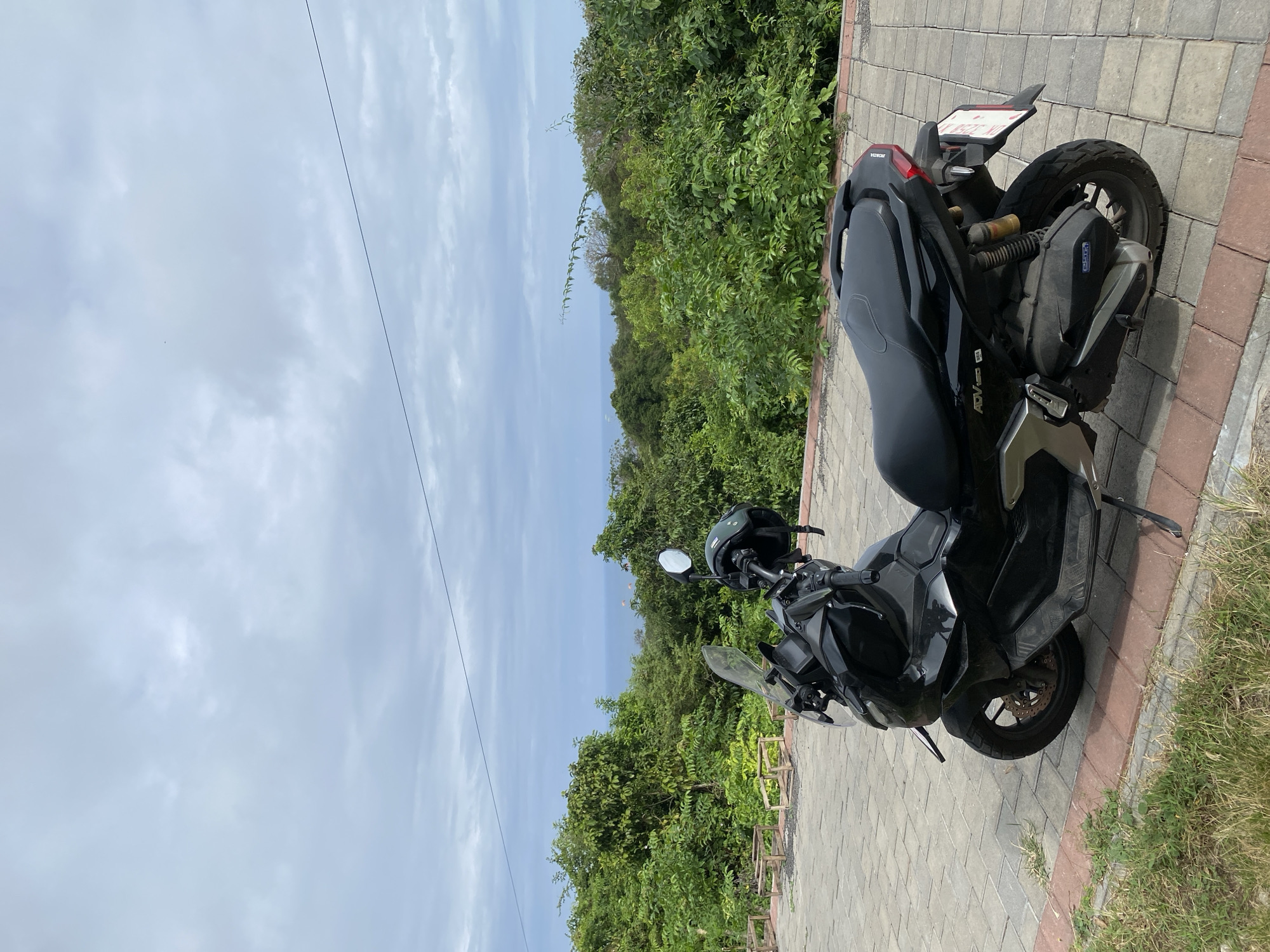 An outdoor, eye-level shot shows a black Honda ADV 150 scooter parked on a paved area with a scenic backdrop. The scooter is the primary subject, angled slightly to the right, revealing its front wheel, handlebars with a black helmet resting on them, and the rear section with a license plate. The paved area is constructed of interlocking gray and red bricks. To the left of the scooter, wooden benches are visible, partially obscured by the vehicle. Beyond the pavement and benches, a lush, green landscape of trees and foliage dominates the midground. In the distance, the faint blue line of the ocean meets a cloudy sky. The overall scene suggests a parking area with a view, likely in Kuta Selatan, Indonesia, given the tropical vegetation and scooter model commonly found in the region. The lighting indicates an overcast day, with diffused natural light. No people are present in the frame.