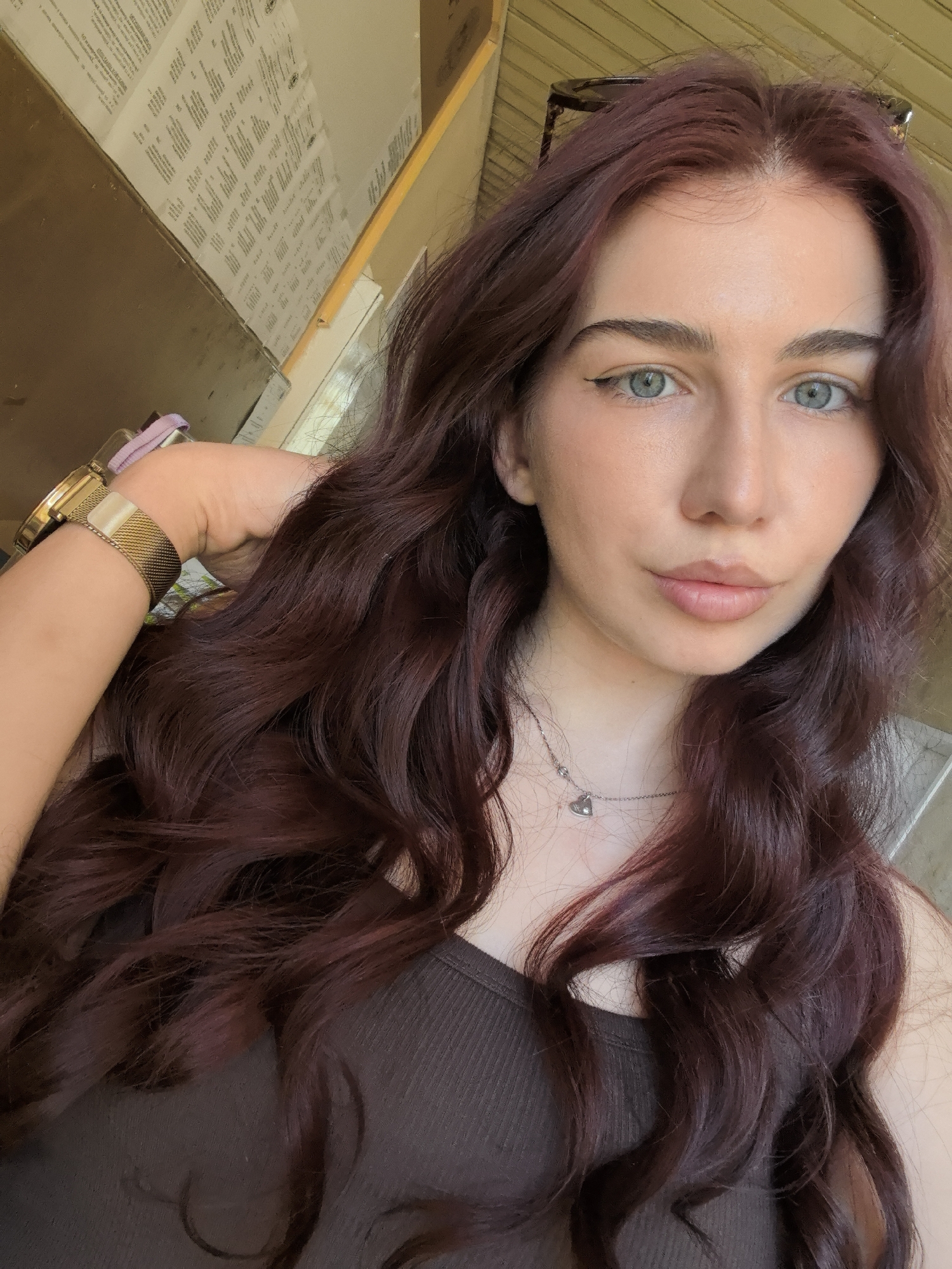 A close-up selfie of a young woman with wavy, reddish-brown hair and light blue eyes. She is wearing a brown tank top and a gold watch. A silver necklace with a heart pendant is visible. The background appears to be an interior space with boxes on a shelf. It is not possible to discern a location in Bogomilovo, Bulgaria, from this image.